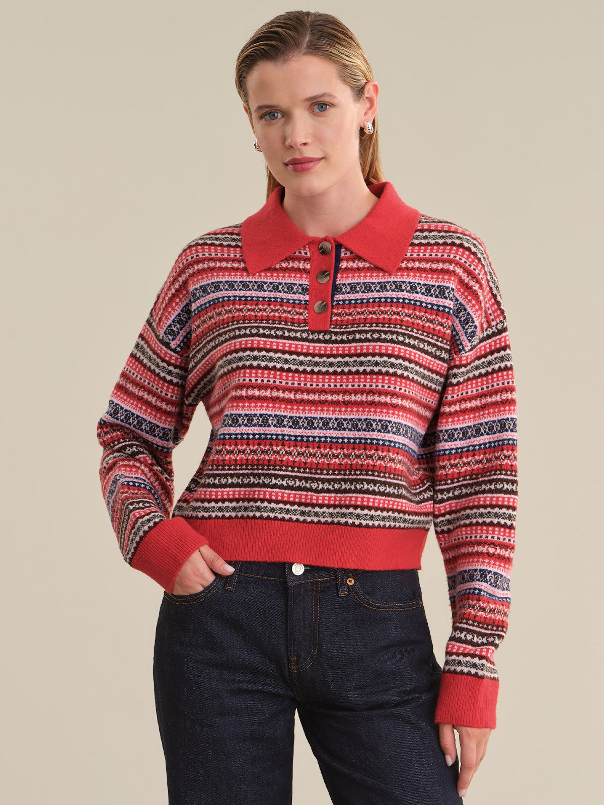 A woman modeling a cropped knit polo sweater with a red collar, cuffs, and hem, featuring horizontal multicolor Fair Isle stripes in red, navy, cream, and black, paired with dark blue jeans against a solid beige background.