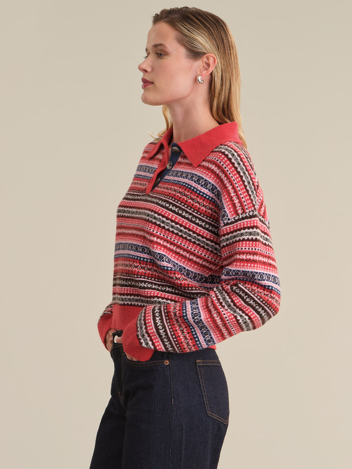 A woman shown in side profile wearing a long-sleeve knit polo-style sweater with horizontal multicolor stripes in red, navy, black, and white, paired with dark blue jeans, against a solid beige studio background.