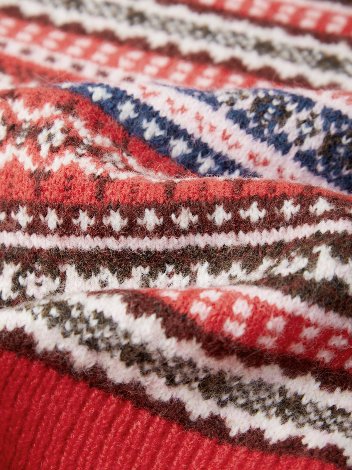 A close-up of a knitted fabric with a Fair Isle pattern in red, white, blue, and brown, showing the texture and detailed knit stitches.