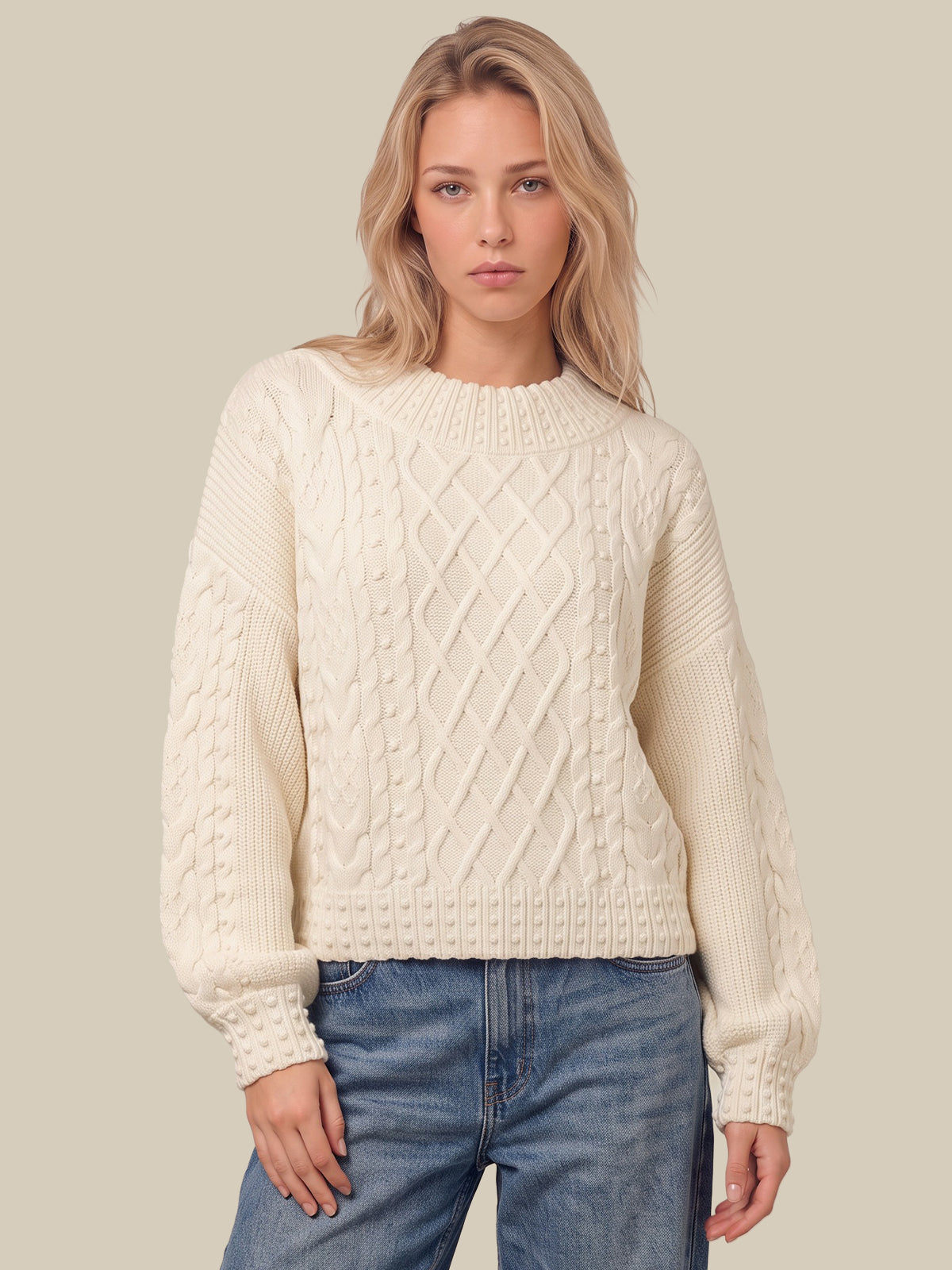 A blonde woman wearing a cream-colored cable-knit sweater and blue jeans, standing against a solid beige background.
