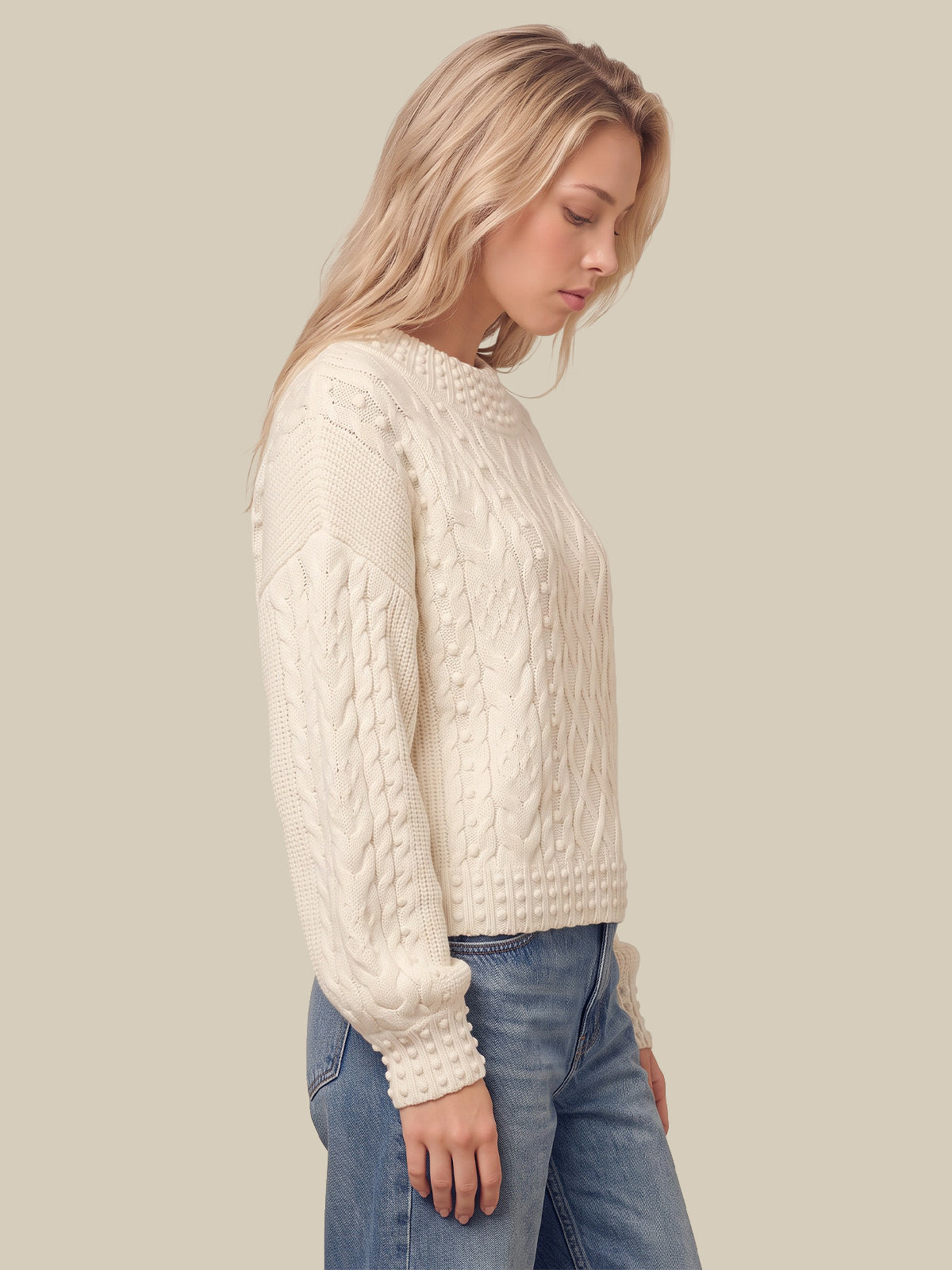 A blonde woman wearing a cream cable-knit sweater and blue jeans, shown in profile against a solid light beige background.