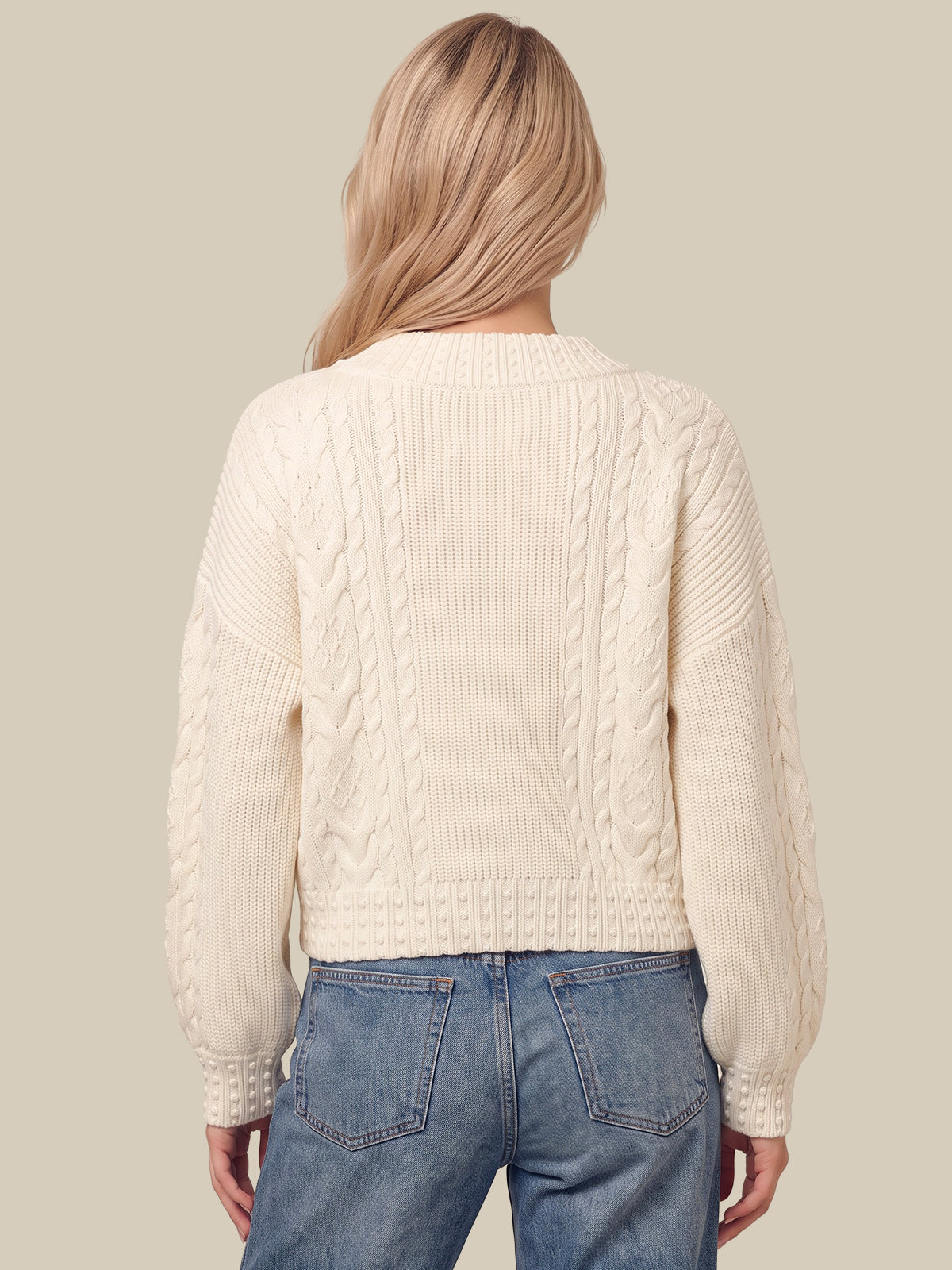 A woman viewed from the back wearing a cream cable-knit sweater and blue jeans against a solid beige background.