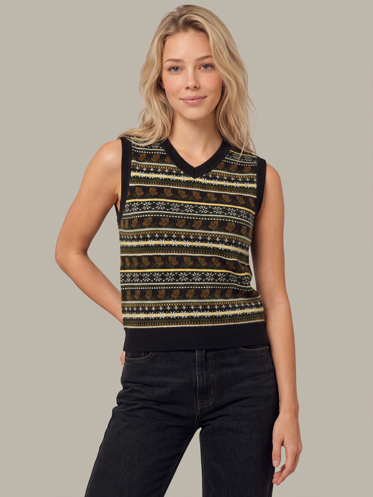 A woman modeling a sleeveless knit sweater vest with a black V-neck and horizontal Fair Isle style stripes in brown, beige, and black, paired with dark jeans against a solid gray studio background.