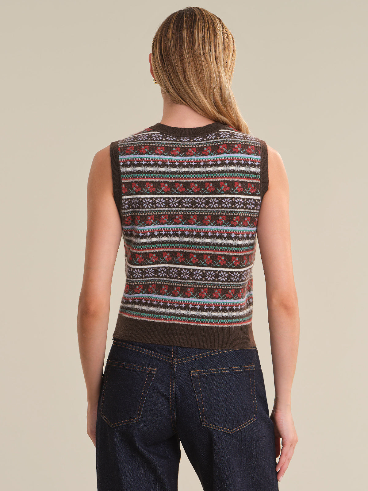 A woman viewed from the back wearing a sleeveless knit vest with horizontal multicolor Fair Isle-style patterns in red, white, blue, and dark brown, paired with dark blue jeans against a plain beige studio background.