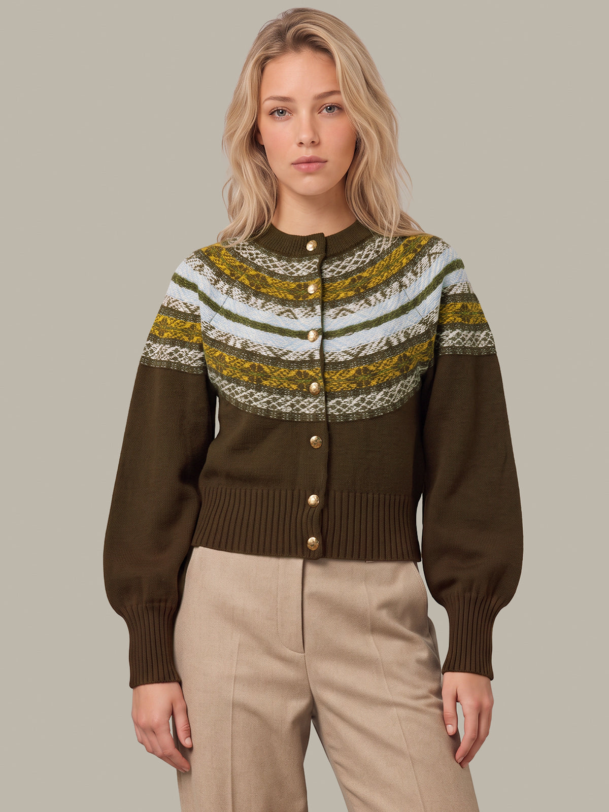 A woman modeling a cropped button-front cardigan with a dark brown base and a multicolor knit yoke featuring olive green, mustard yellow, and cream Fair Isle patterns, paired with beige trousers against a solid gray background.