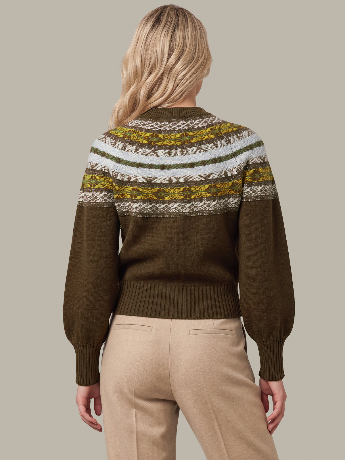 A woman shown from the back wearing a brown knit sweater with a colorful patterned yoke in olive green, cream, and mustard yellow, paired with beige trousers, against a solid taupe background.