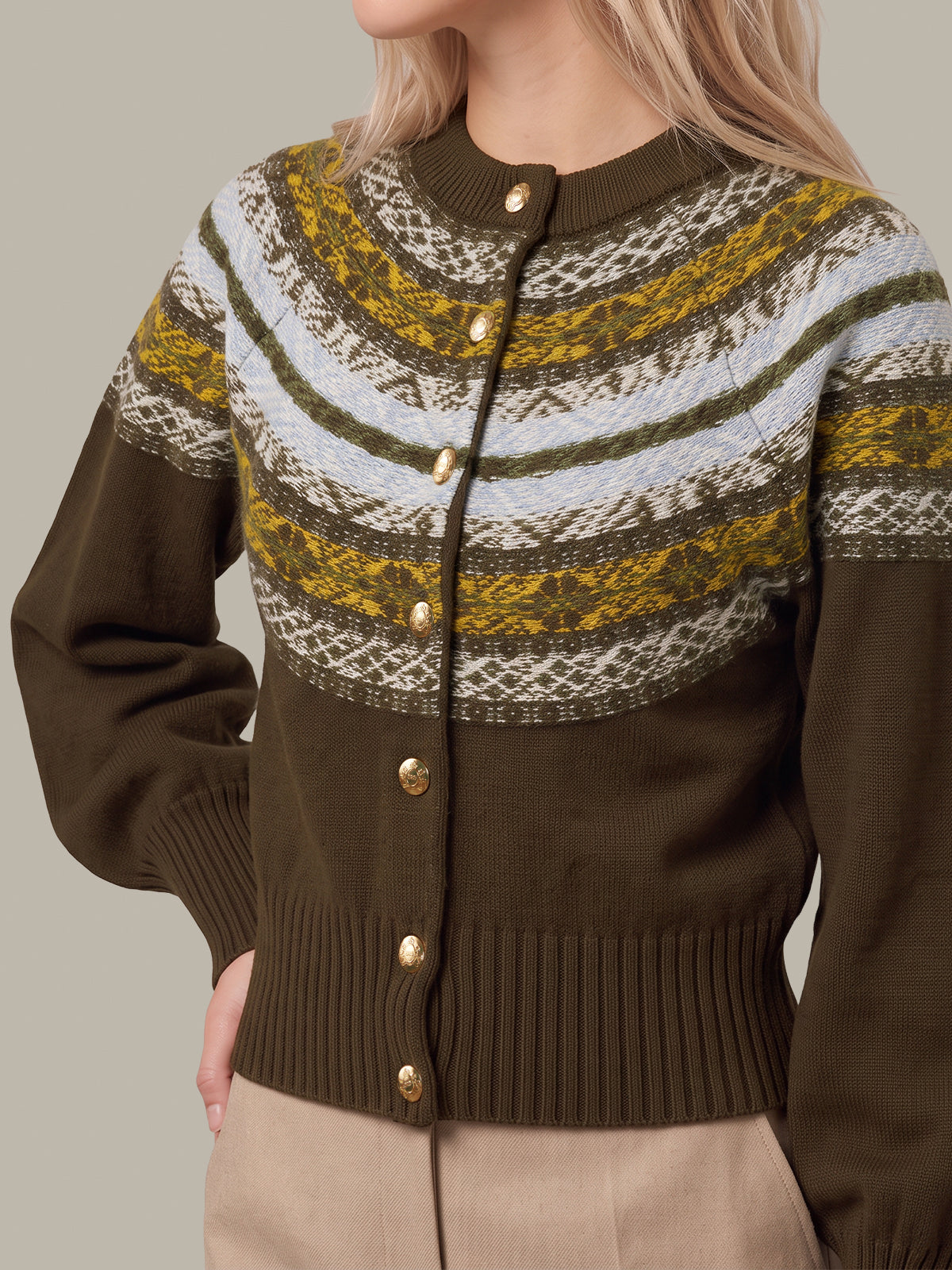 A close-up of a woman wearing a brown knit cardigan sweater with a colorful Fair Isle pattern across the chest and shoulders, featuring gold buttons and beige pants, set against a solid gray background.
