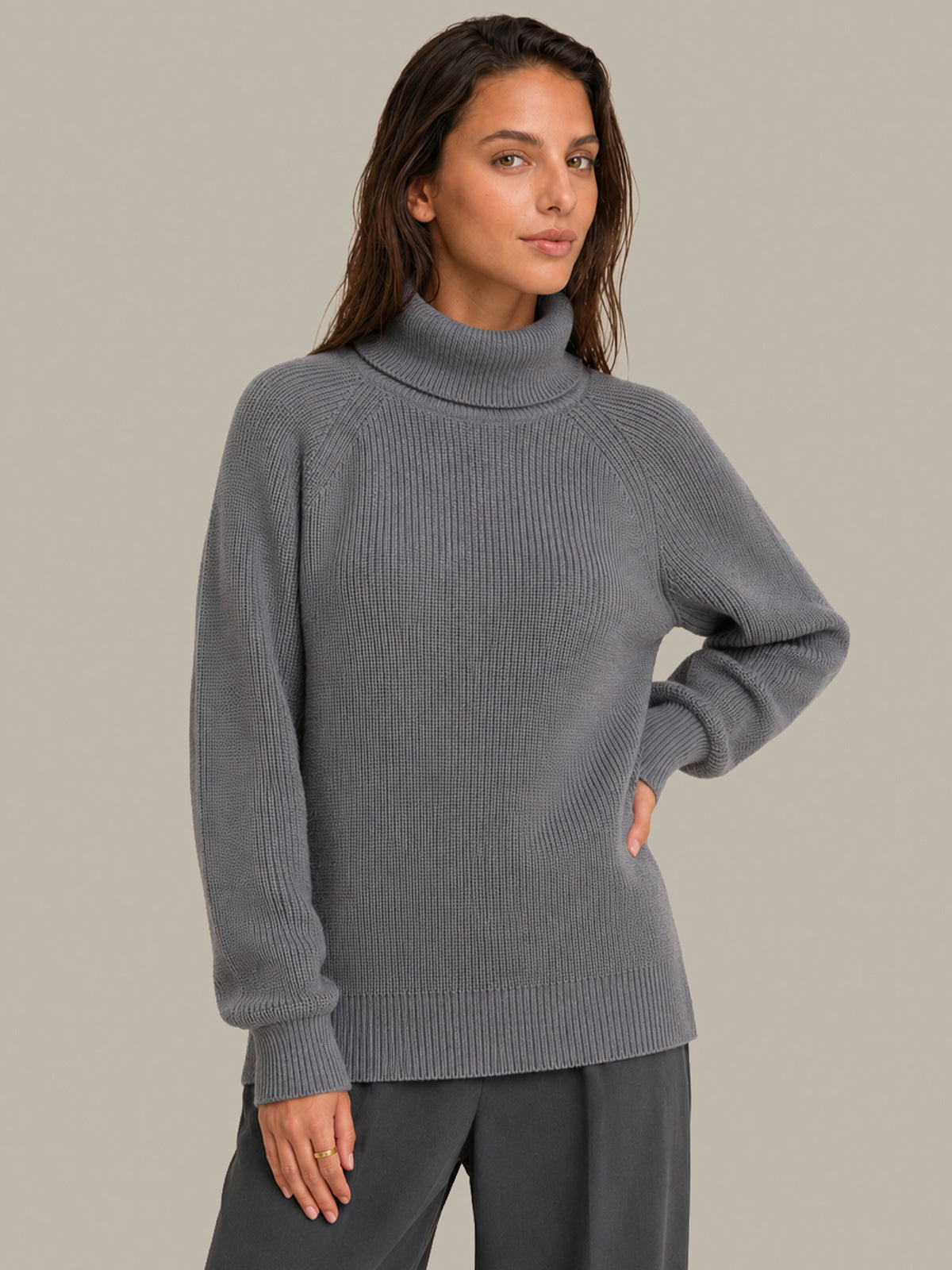 A woman wearing a ribbed gray mock neck sweater and dark gray pants, standing against a solid light gray background.