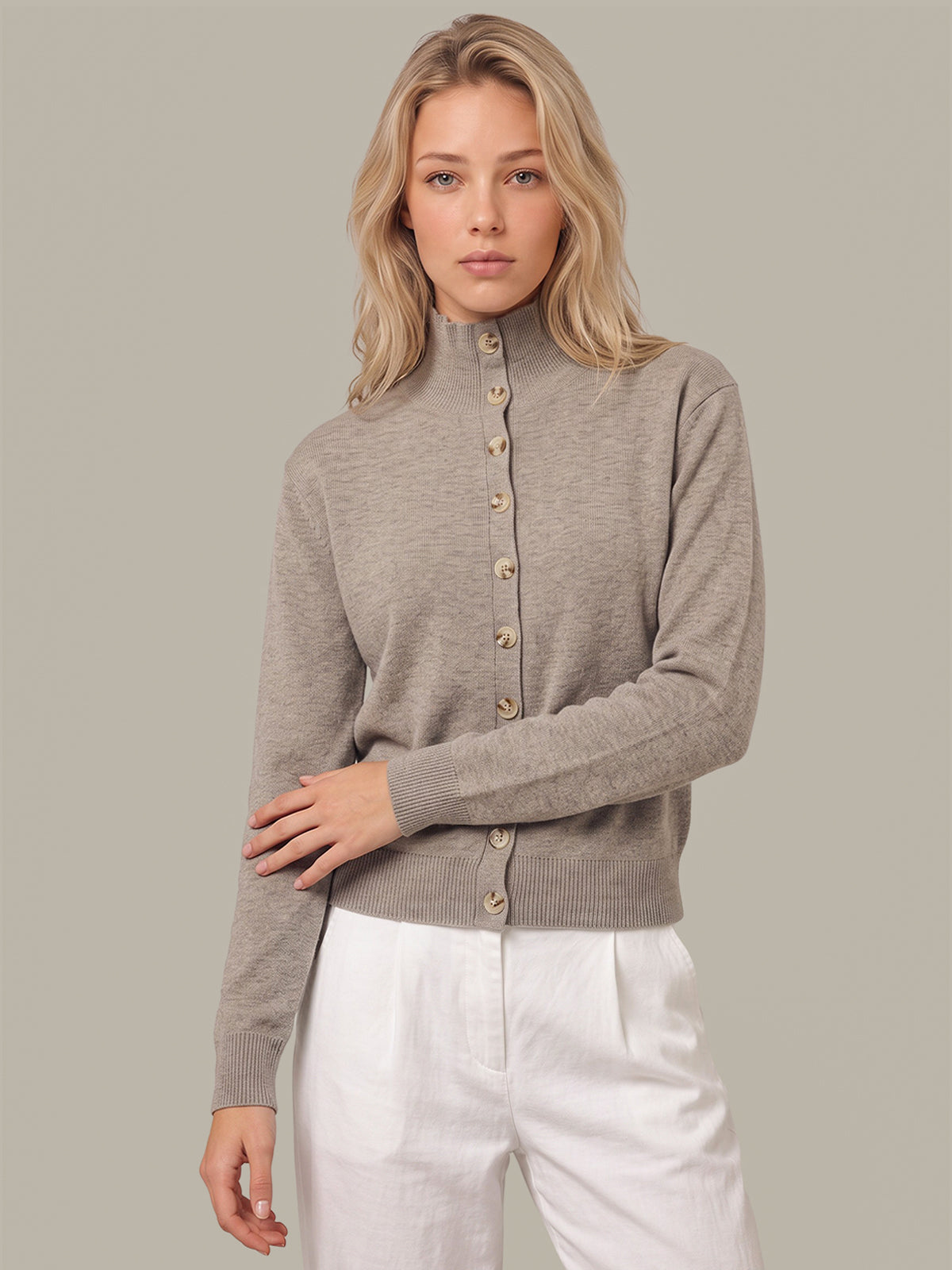 A blonde woman wearing a taupe-gray mock neck button-up cardigan sweater and white trousers, standing against a solid gray studio background.