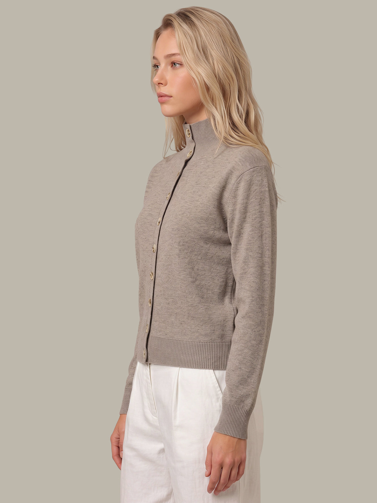 A woman shown in side profile wearing a taupe-gray mock neck cardigan with gold buttons and white trousers, set against a solid light gray background.