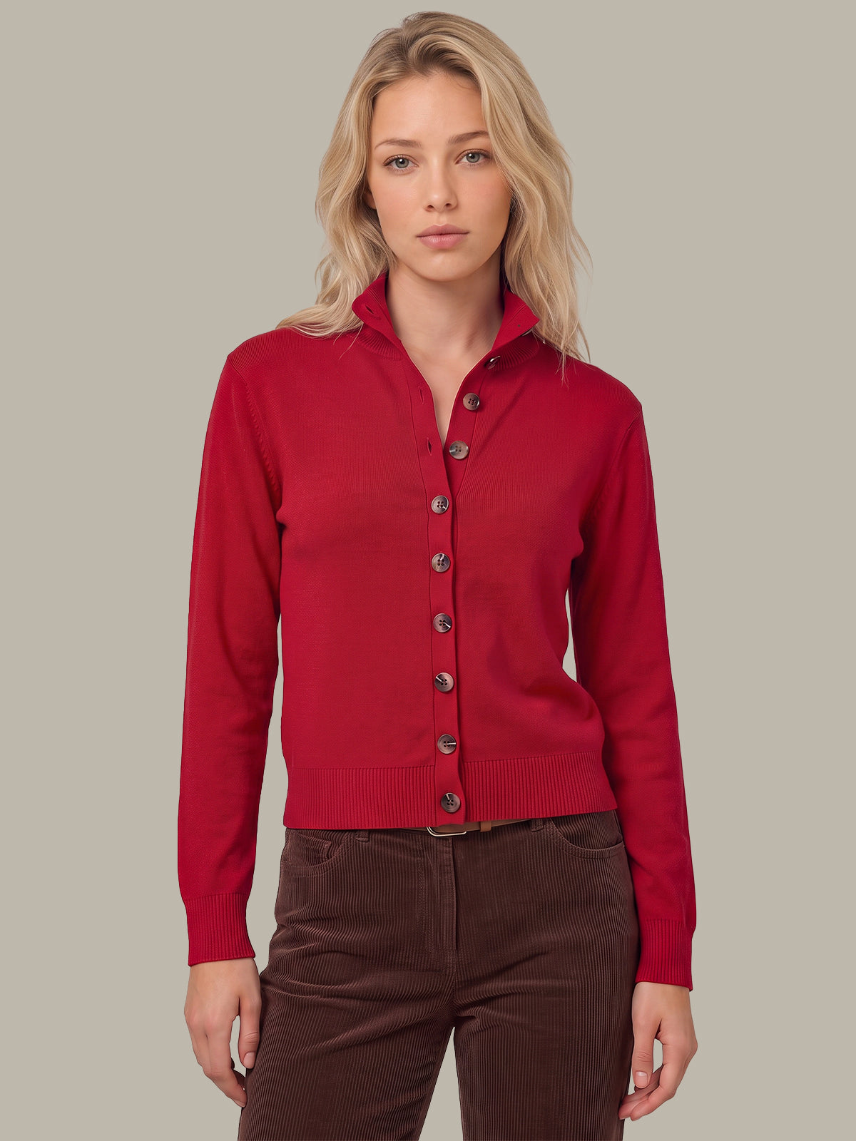 A blonde woman wearing a red button-up knit cardigan with a stand collar and dark brown pants, standing against a solid light gray background.