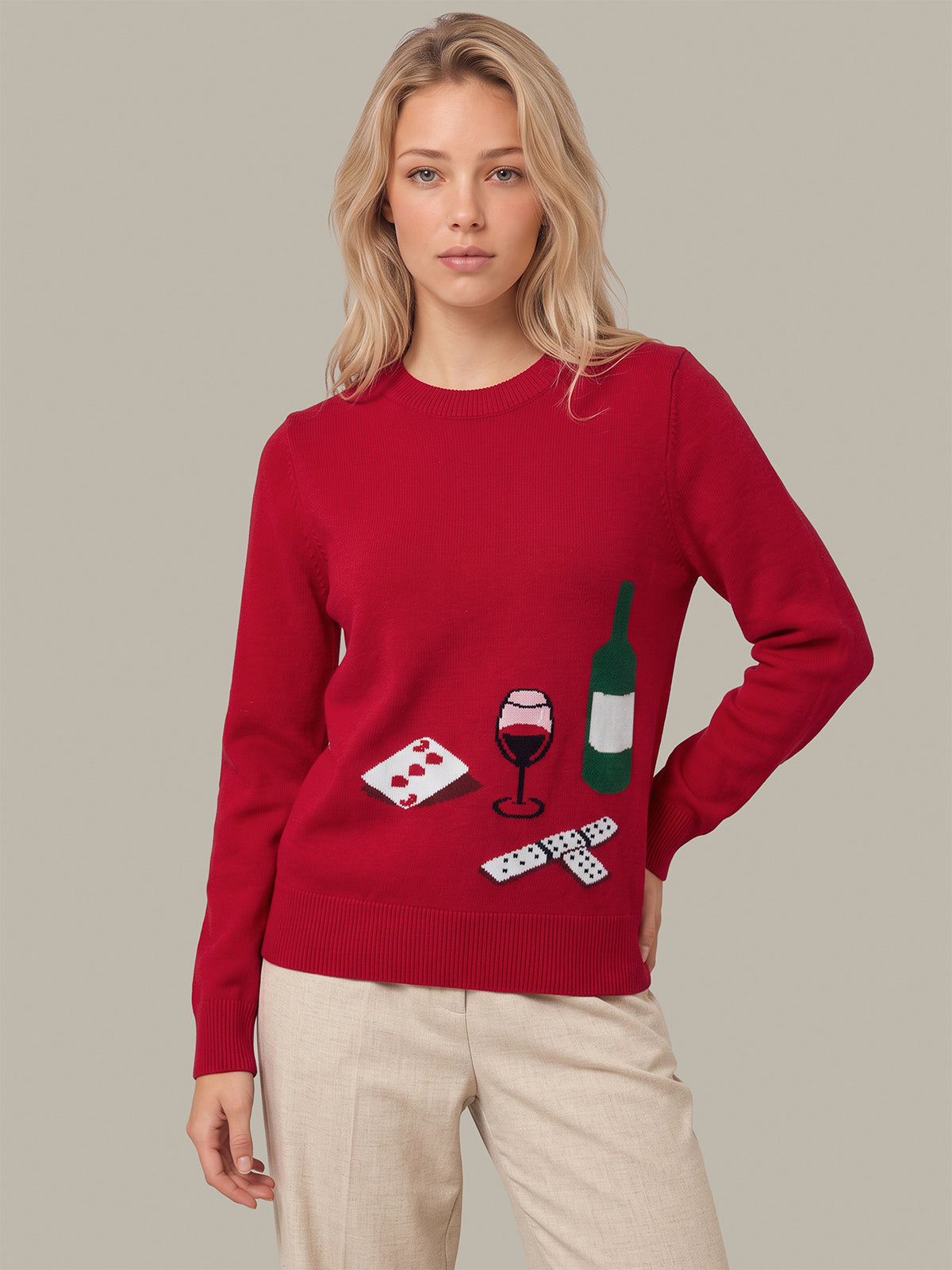 A woman modeling a red crewneck sweater with a graphic print of a wine bottle, wine glass, playing cards, and dominoes, paired with beige trousers, against a solid gray background.