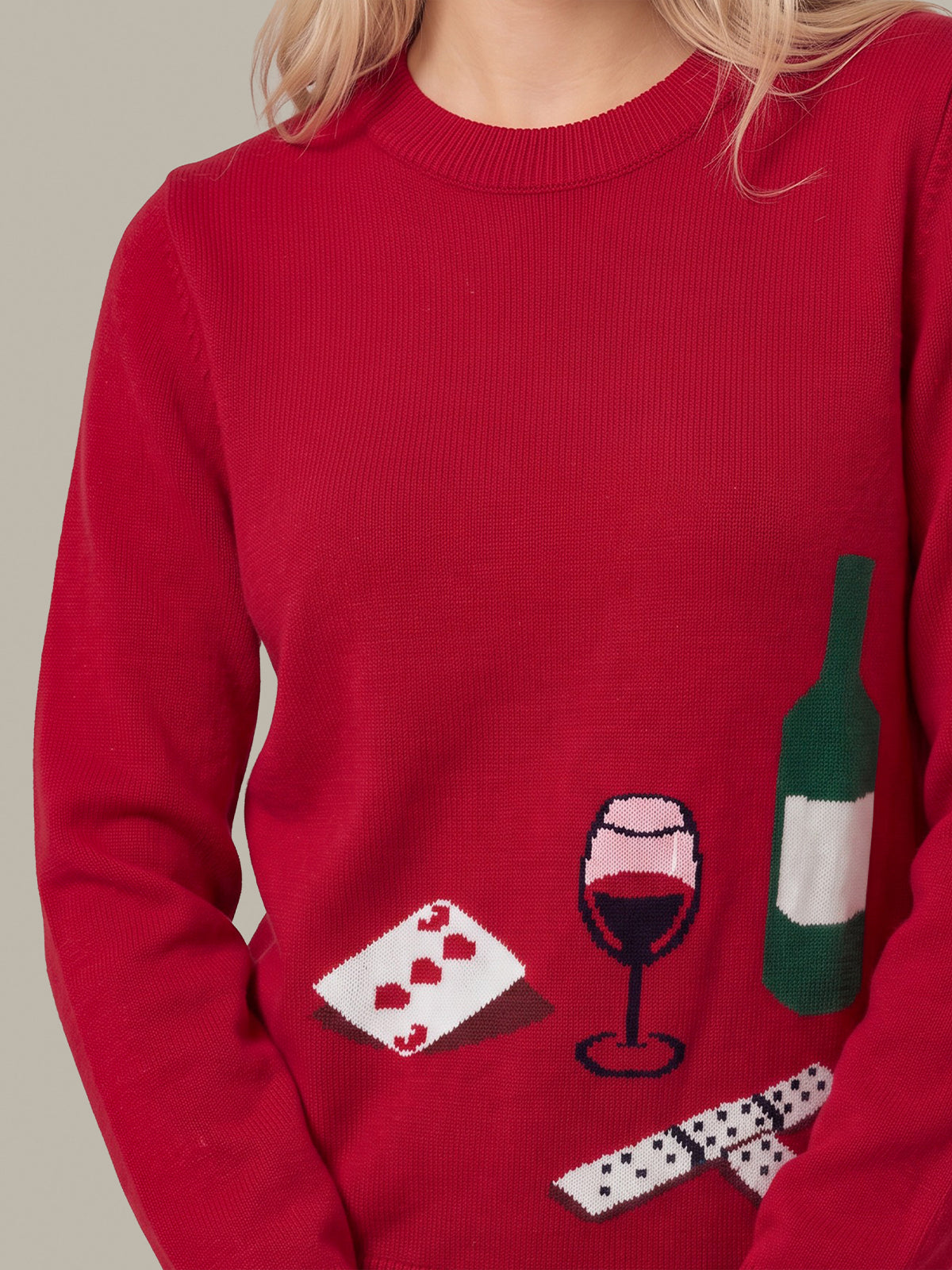 A close-up of a person wearing a bright red knit sweater with a festive wine-themed graphic on the front, featuring a playing card, wine glass, bottle, and dominoes.