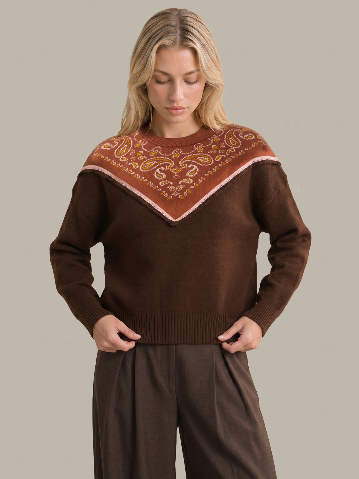 A woman wearing a brown knit sweater with a wide collar featuring a rust-orange and beige paisley pattern, paired with brown trousers, against a solid light gray background.