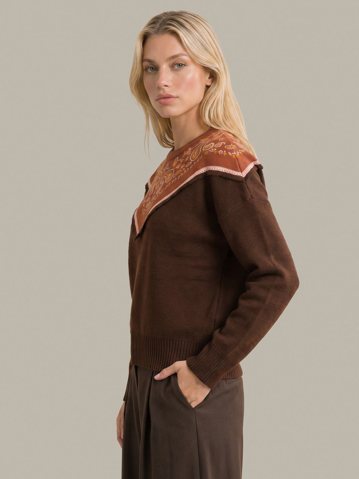 A blonde woman wearing a brown knit sweater with a contrasting patterned yoke in rust orange, beige, and white, paired with dark brown trousers, set against a solid light gray background.