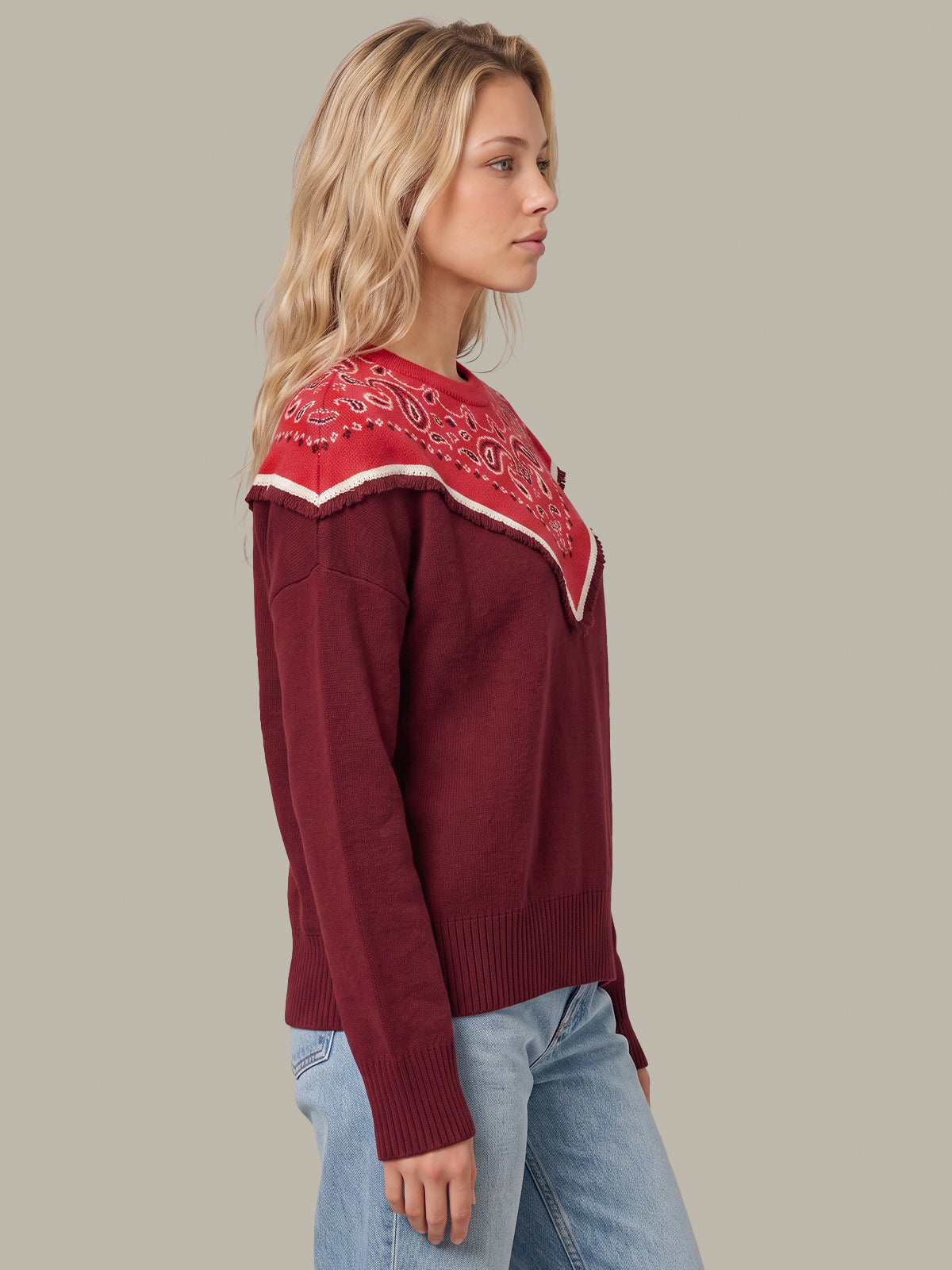 A woman wearing a burgundy knit sweater with a red and white paisley bandana collar, paired with light blue jeans, shown from a side profile against a solid gray background.
