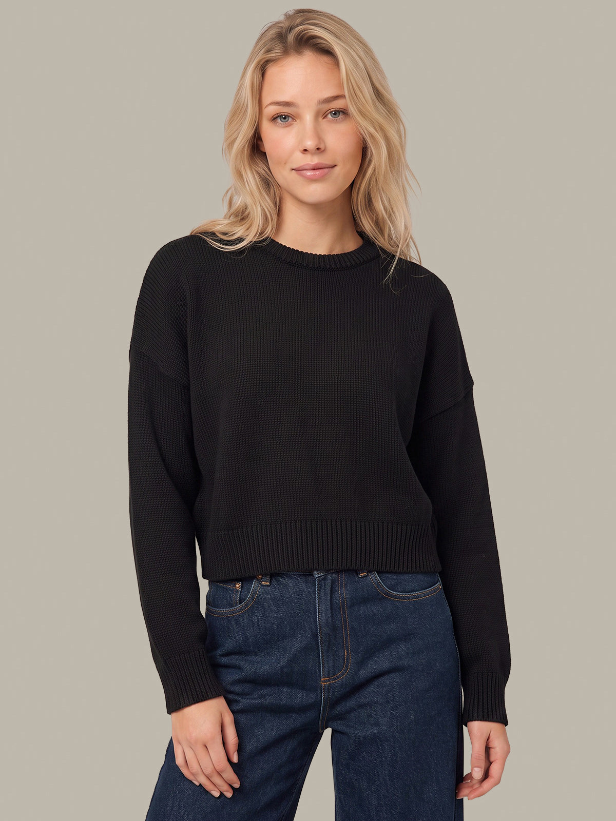 A woman wearing a black ribbed knit crewneck sweater and dark blue jeans, standing against a solid light gray background.