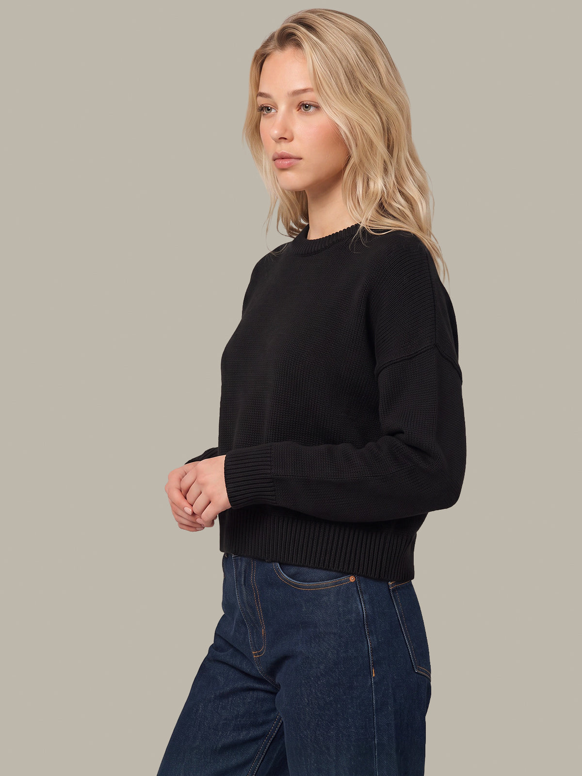 A woman wearing a black long-sleeve knit sweater and dark blue jeans, photographed against a solid light gray studio background.