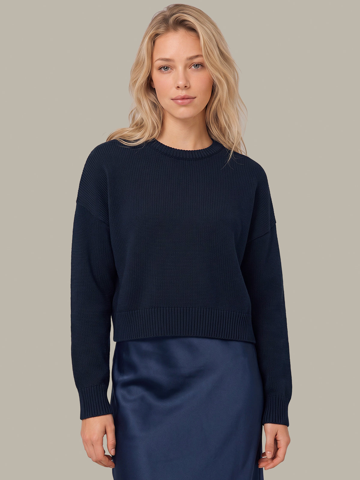 A blonde woman wearing a dark navy ribbed knit crewneck sweater and a matching navy satin skirt, standing against a solid light gray background.