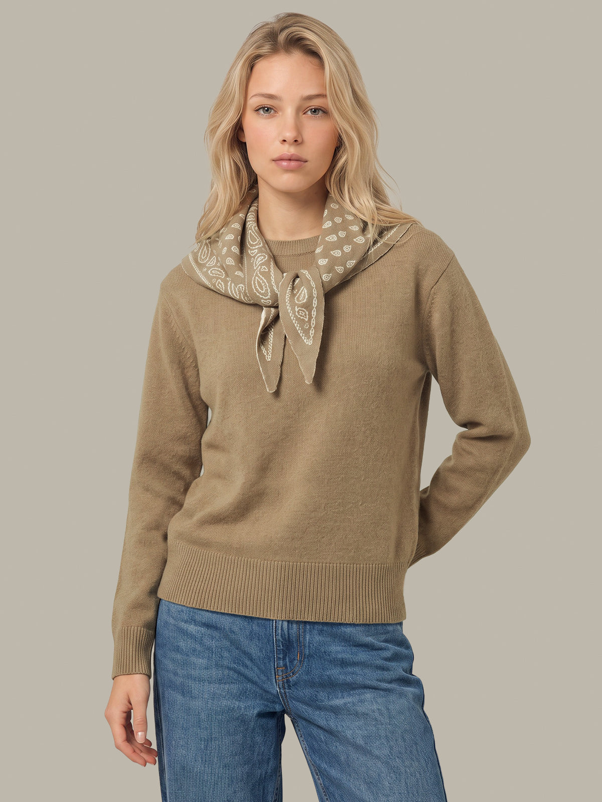 A woman modeling a tan knit sweater with a matching patterned neck scarf, paired with blue jeans, against a solid gray studio background.