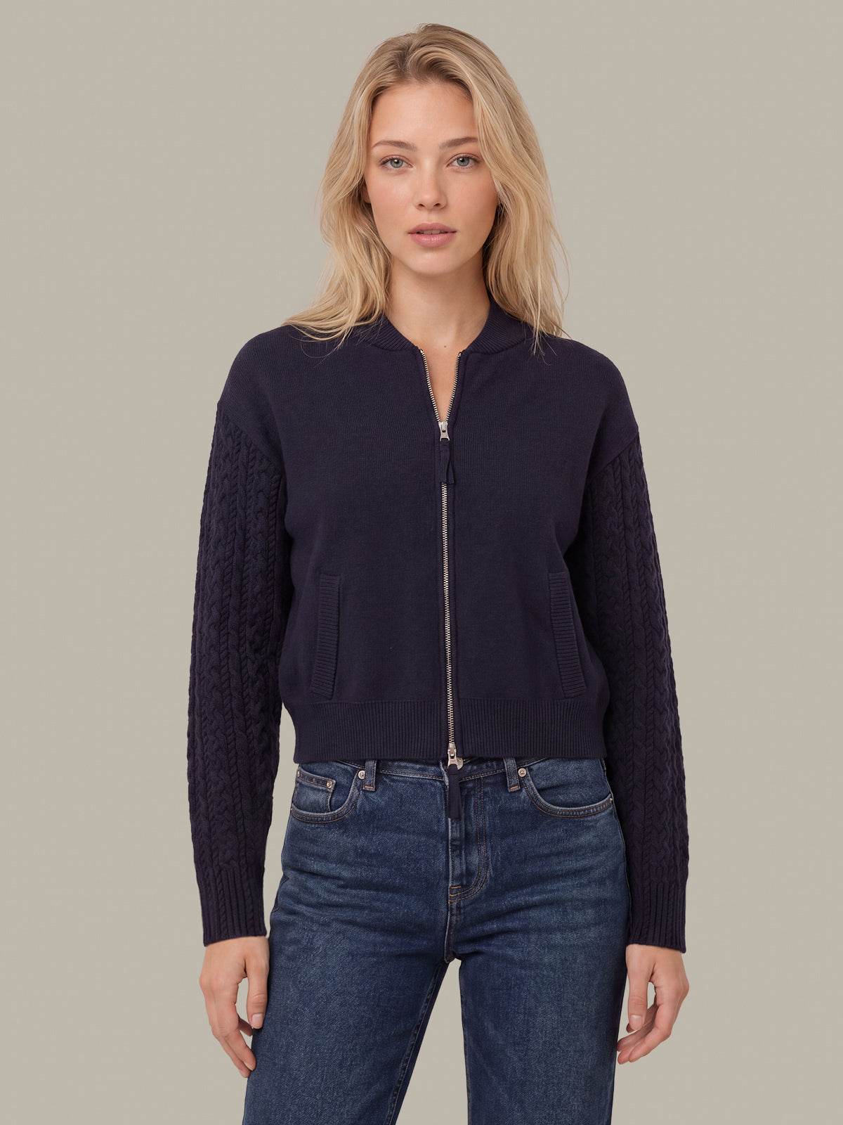 A woman modeling a navy blue zip-up knit jacket with textured cable-knit sleeves, paired with dark blue jeans, against a solid beige background.