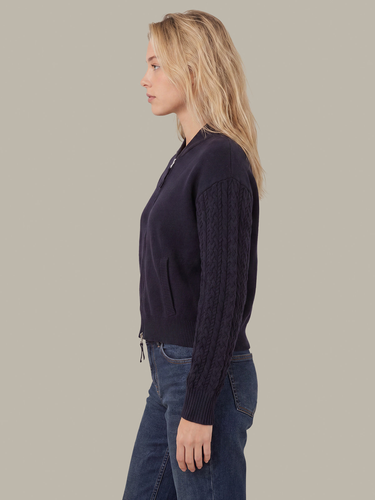 A woman shown in side profile wearing a dark navy cable-knit zip-front cardigan with ribbed trim, paired with blue jeans, against a solid light gray background.