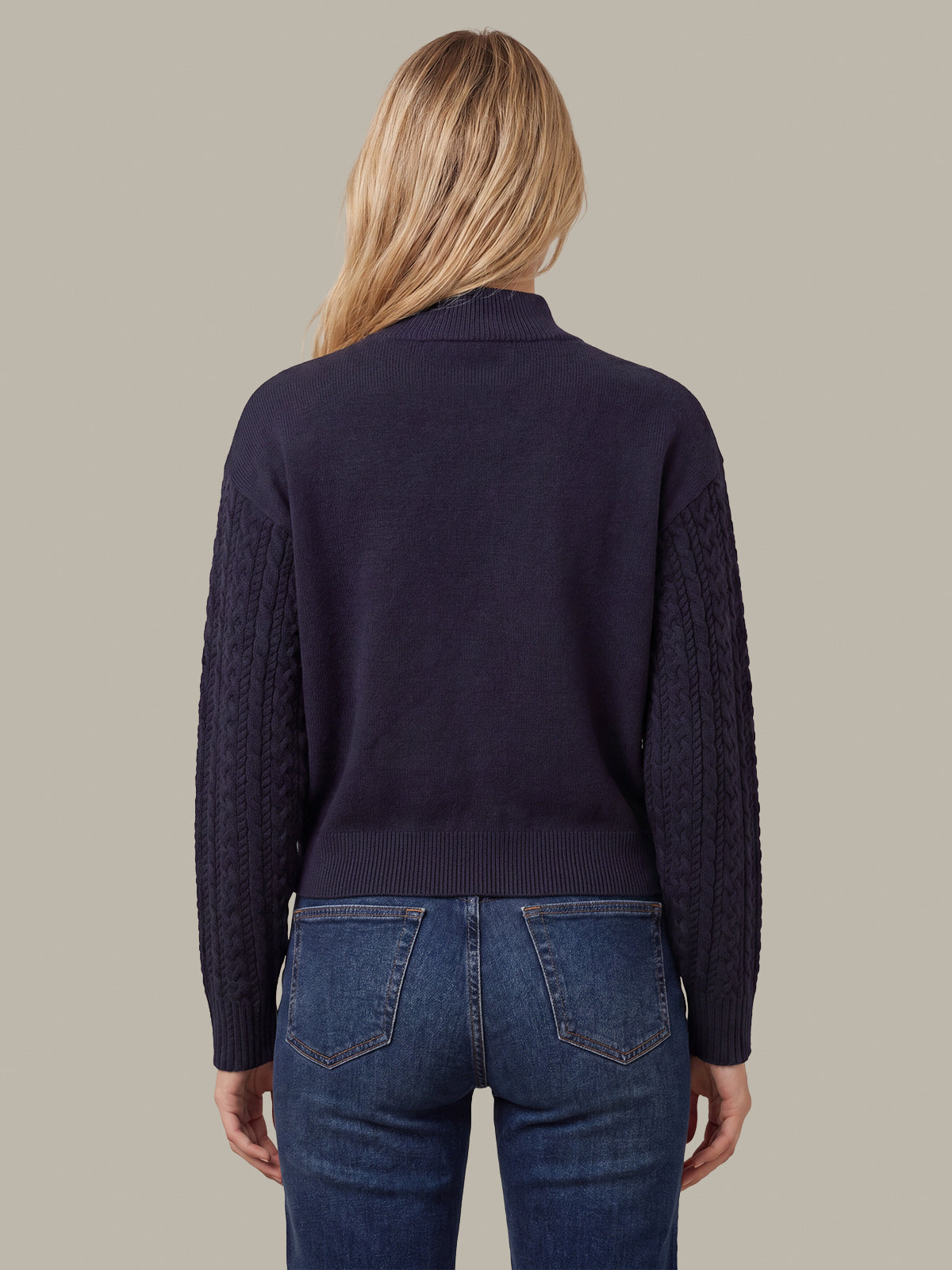 A woman viewed from the back wearing a dark navy cable-knit sweater with a ribbed mock neck and blue jeans, set against a solid taupe background.