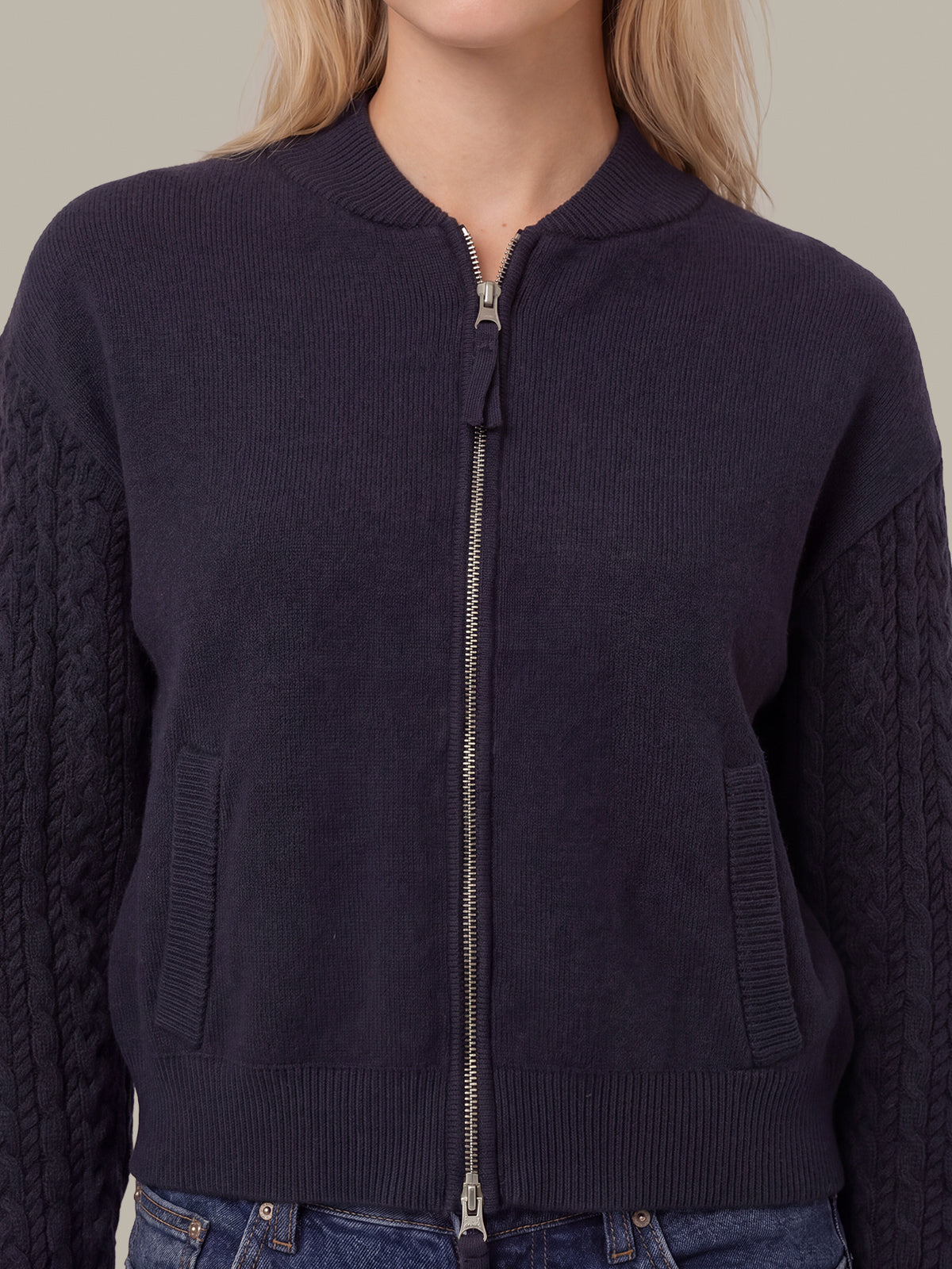 A close-up of a person wearing a dark navy ribbed knit zip-front sweater with cable-knit sleeves and side pockets, paired with blue jeans against a plain beige background.