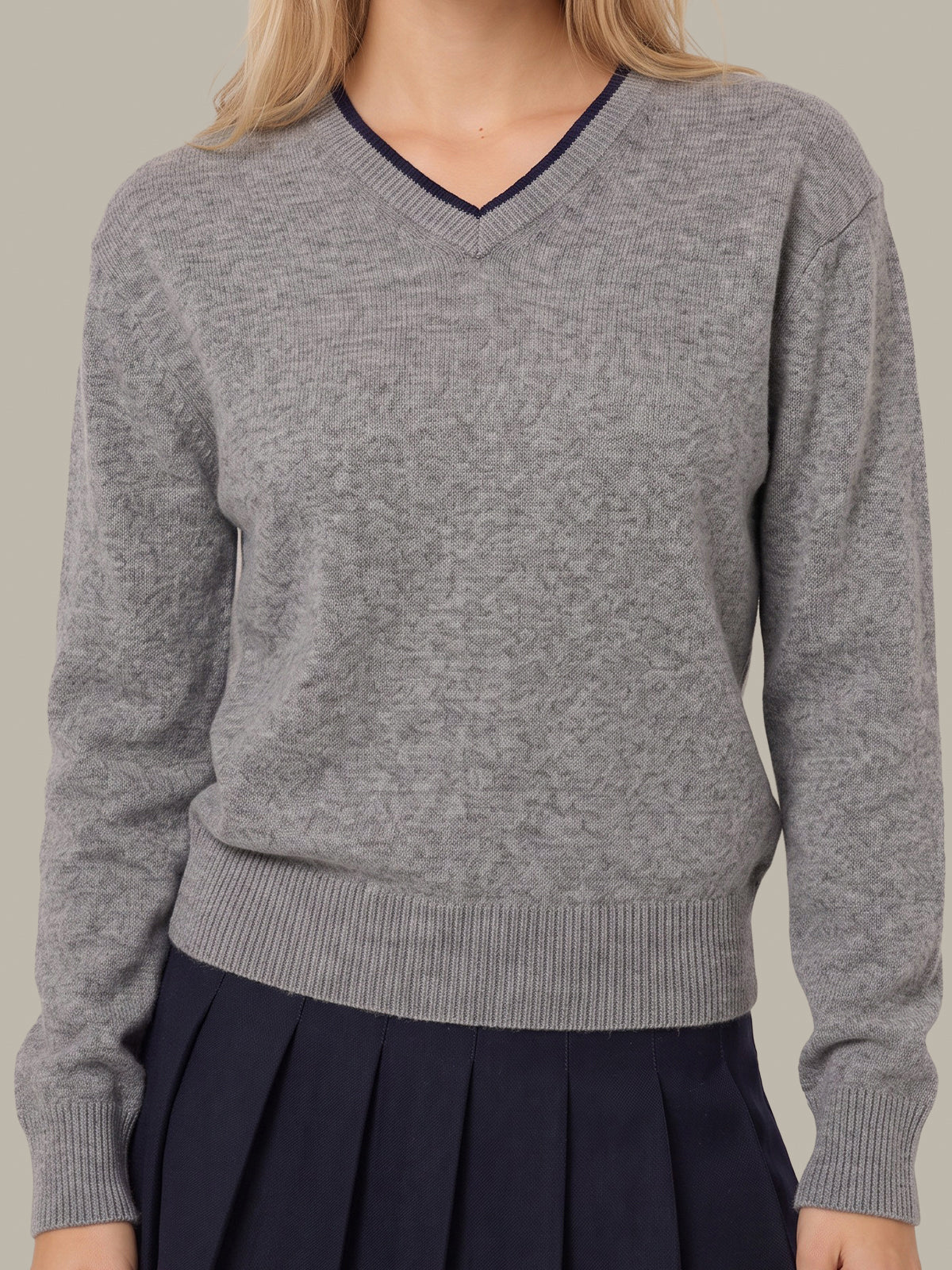 A woman wearing a heather gray long-sleeve V-neck sweater with dark navy trim along the neckline, paired with a dark navy pleated skirt, against a solid light gray background.