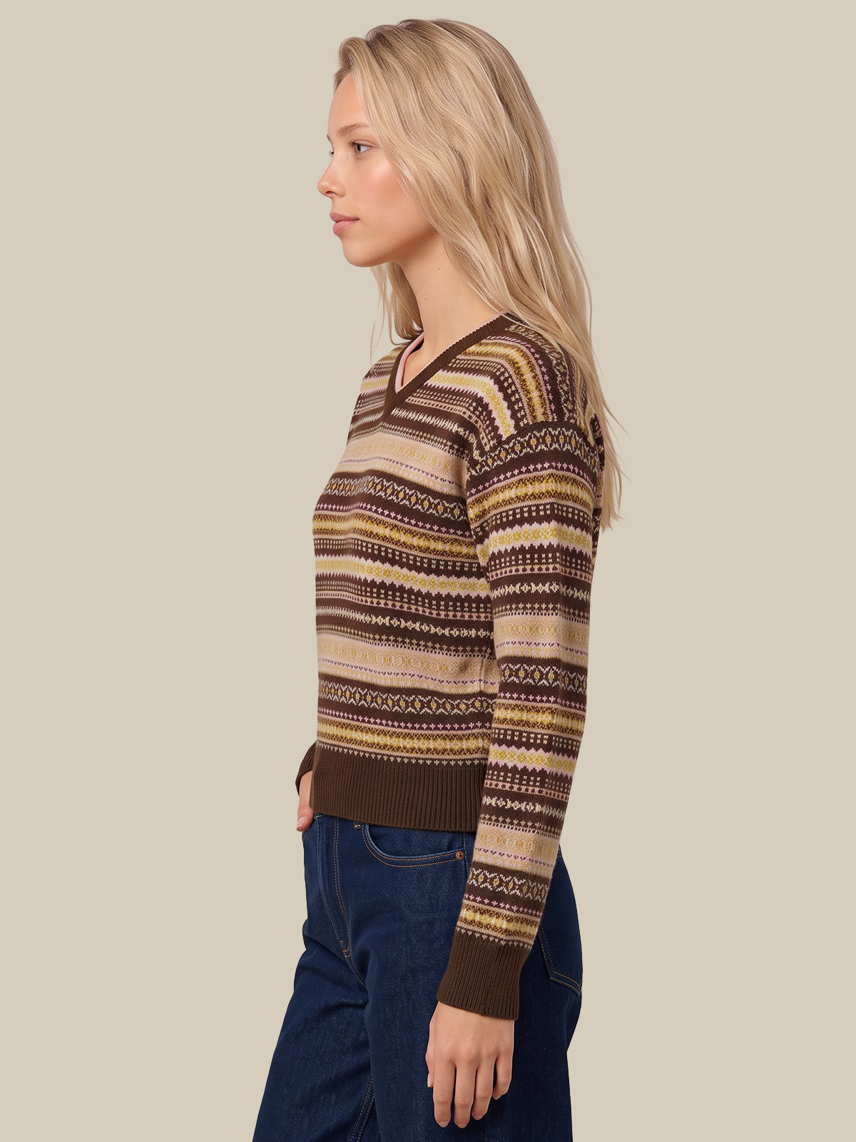 A woman in profile wearing a brown and beige patterned knit sweater with dark blue jeans, set against a solid beige background.