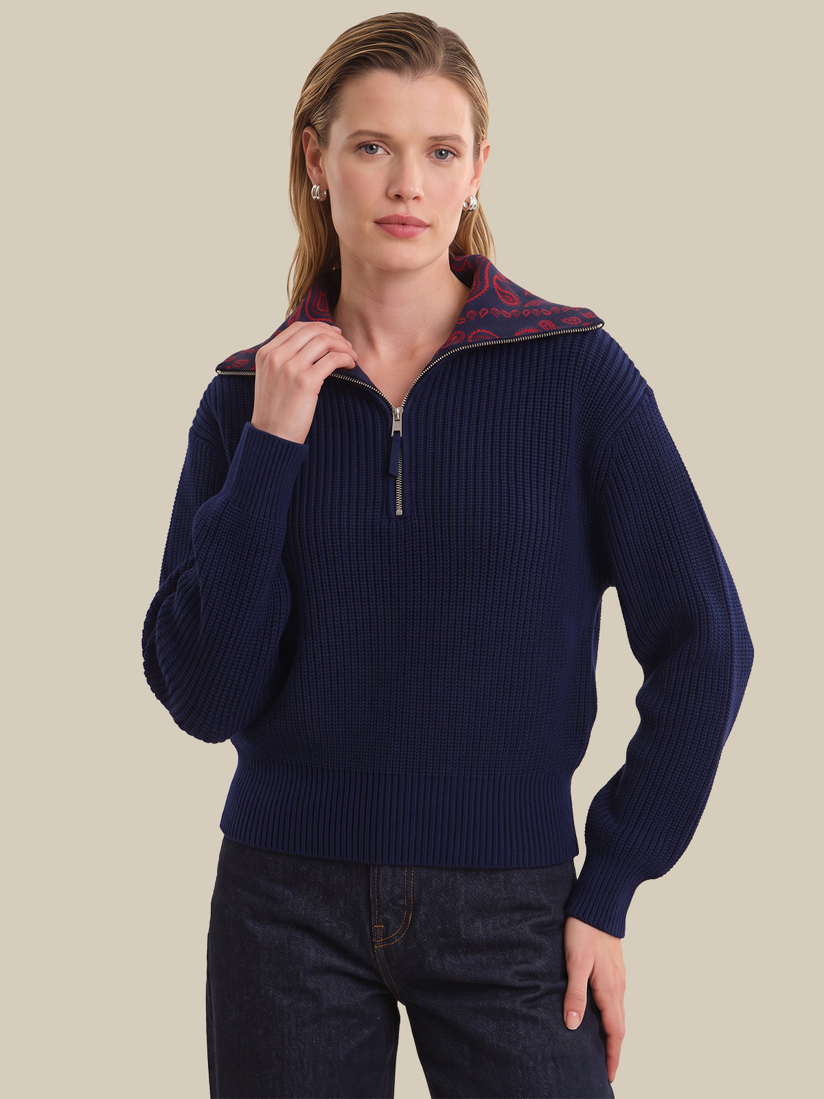 A woman wearing a navy ribbed knit half-zip sweater with a red patterned inner collar, paired with dark blue jeans, against a solid beige background.