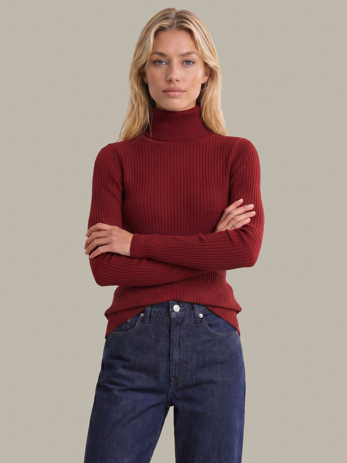 A blonde woman wearing a ribbed mock neck turtleneck sweater in deep burgundy red, paired with dark blue jeans, standing against a solid light gray background.
