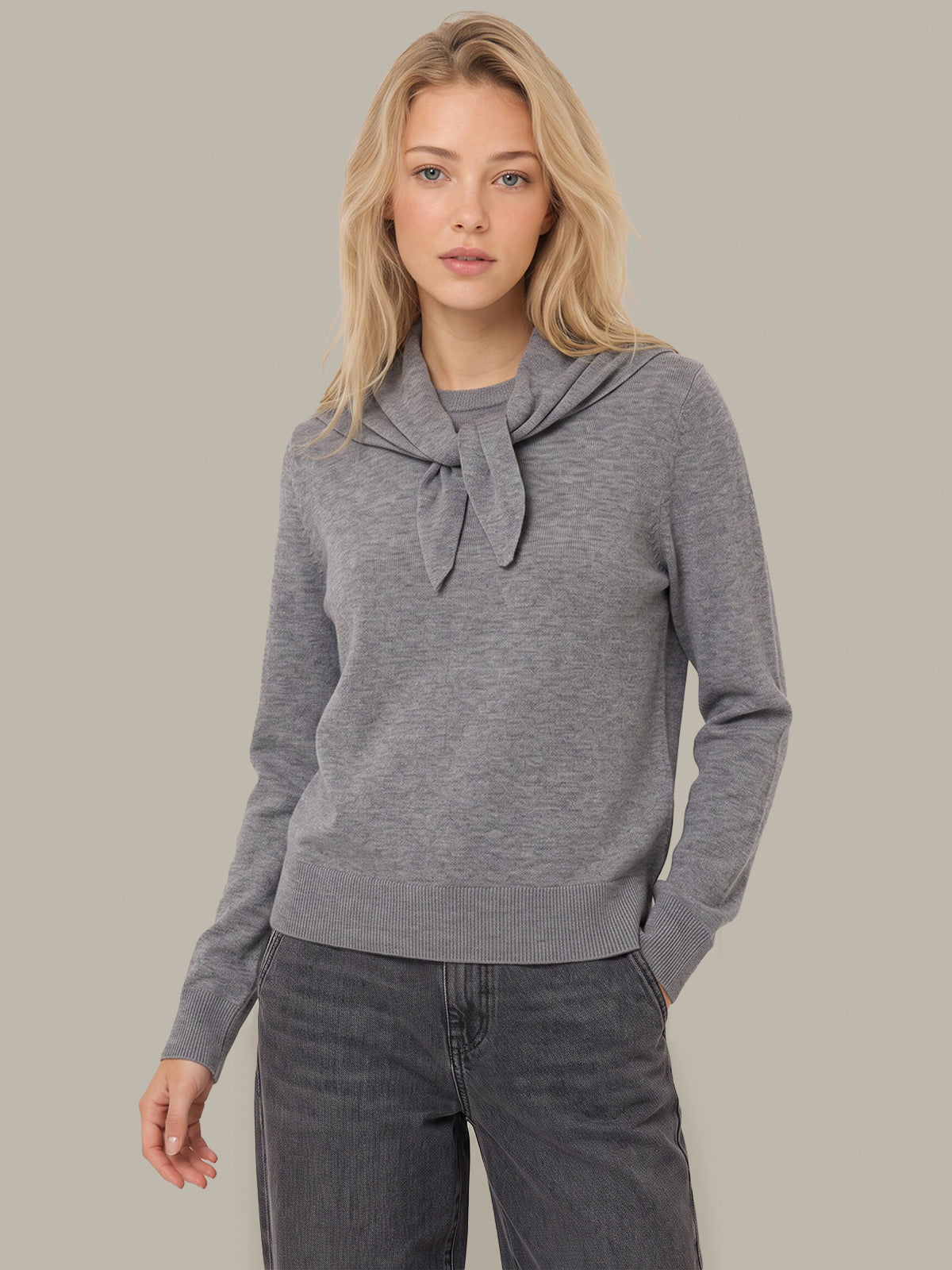 A blonde woman wearing a heather gray long-sleeve knit sweater with a wide draped collar tied in a bow, paired with dark gray jeans, against a solid light gray studio background.