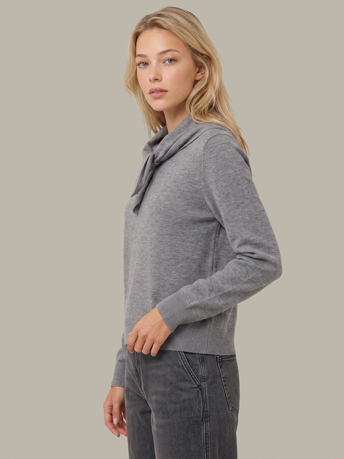 A woman wearing a heather gray long-sleeve knit sweater with a draped cowl neck and dark gray jeans, photographed against a solid warm gray studio background.