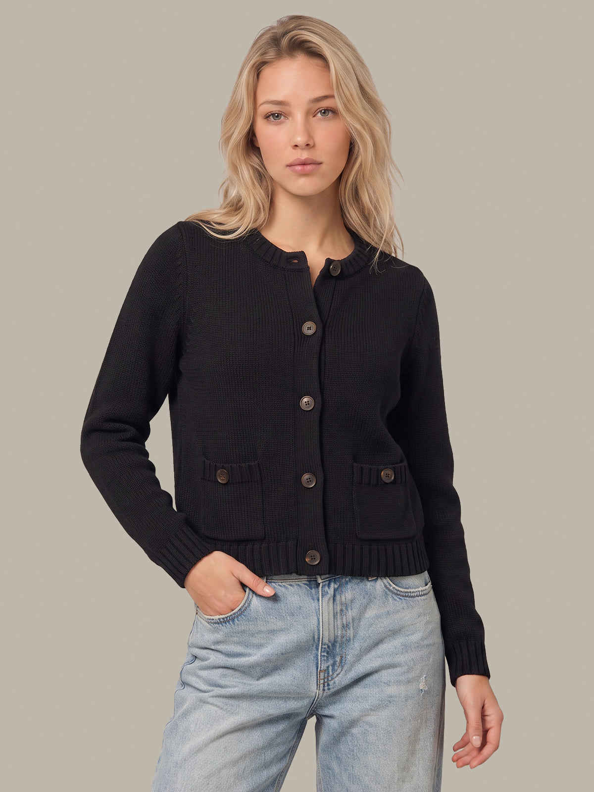 A blonde woman wearing a black ribbed knit cardigan with button closures and front pockets, paired with light-wash blue jeans, against a solid neutral gray background.