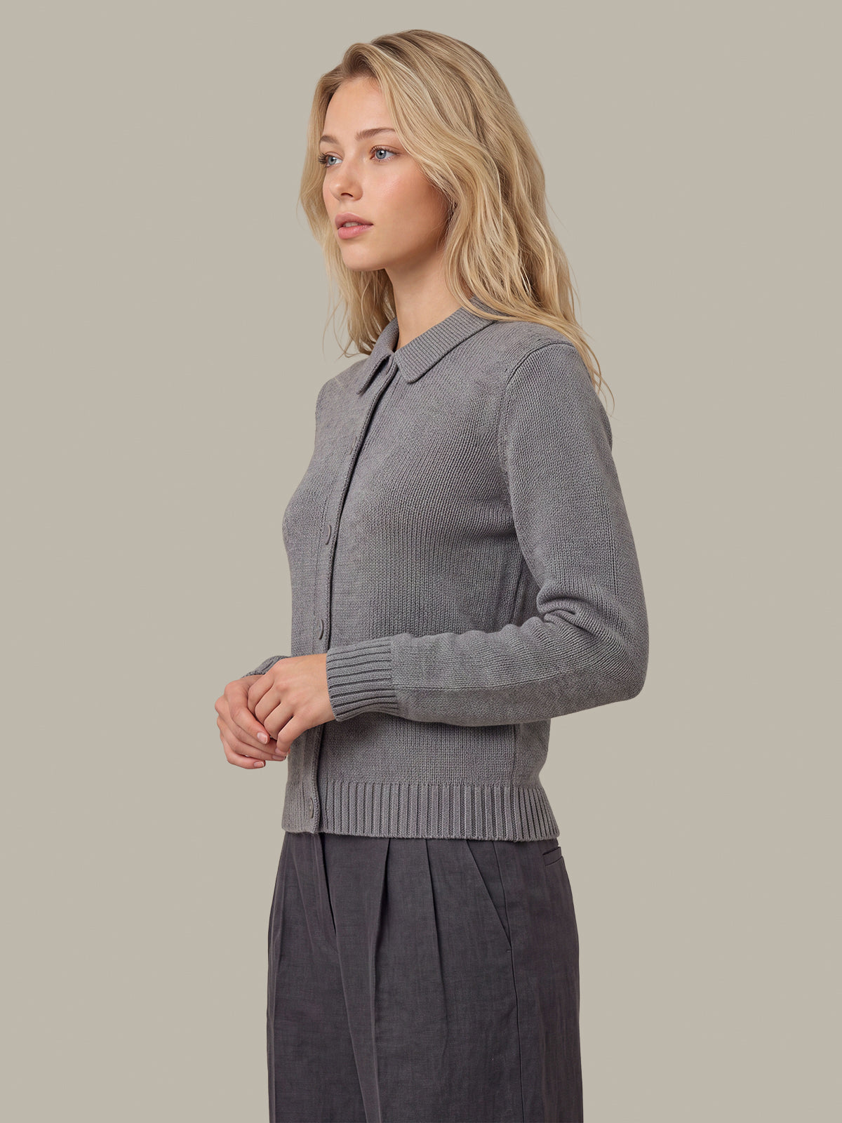 A woman modeling a gray ribbed knit cardigan with a collar and button front, paired with dark gray pleated trousers, against a solid light gray background.