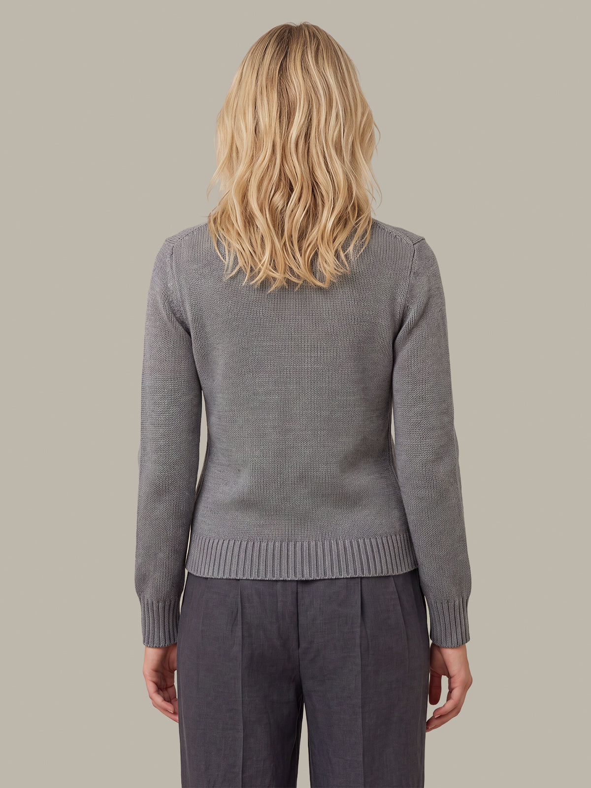 A woman viewed from the back wearing a gray ribbed knit sweater and dark gray trousers, against a solid light gray background.