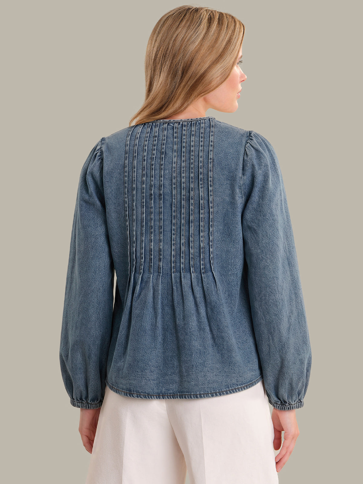 Woman wearing a blue denim blouse with long sleeves and a pleated back on a gray background