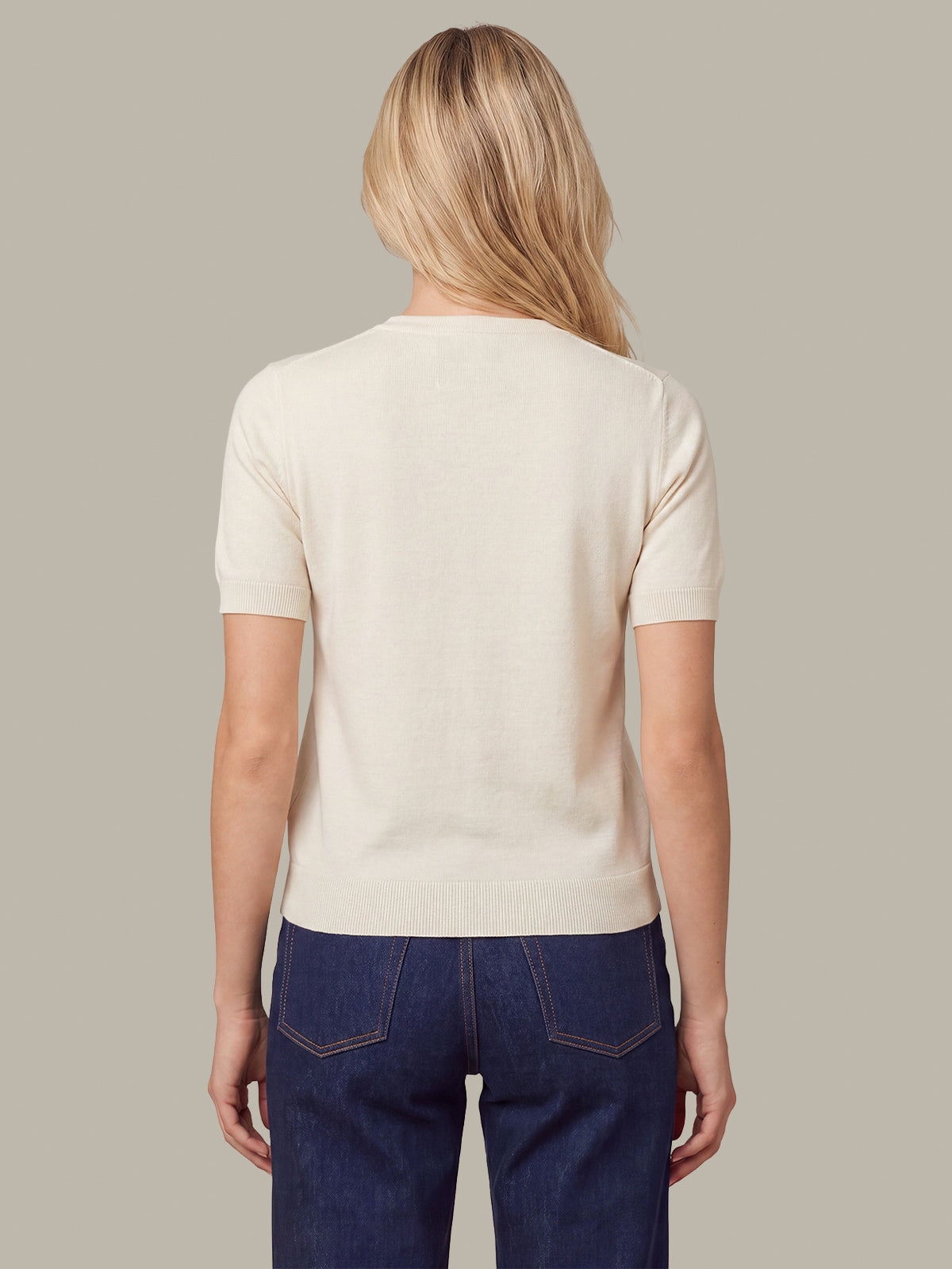 A woman viewed from the back wearing a short-sleeve cream knit top and dark blue jeans, set against a solid light gray background.