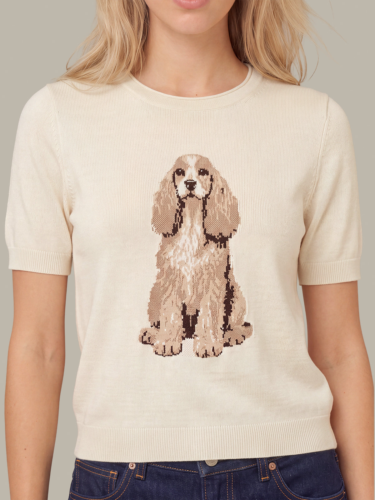 A woman wearing a cream-colored short-sleeve knit sweater with a large beige and brown dog graphic on the front, paired with dark blue jeans.