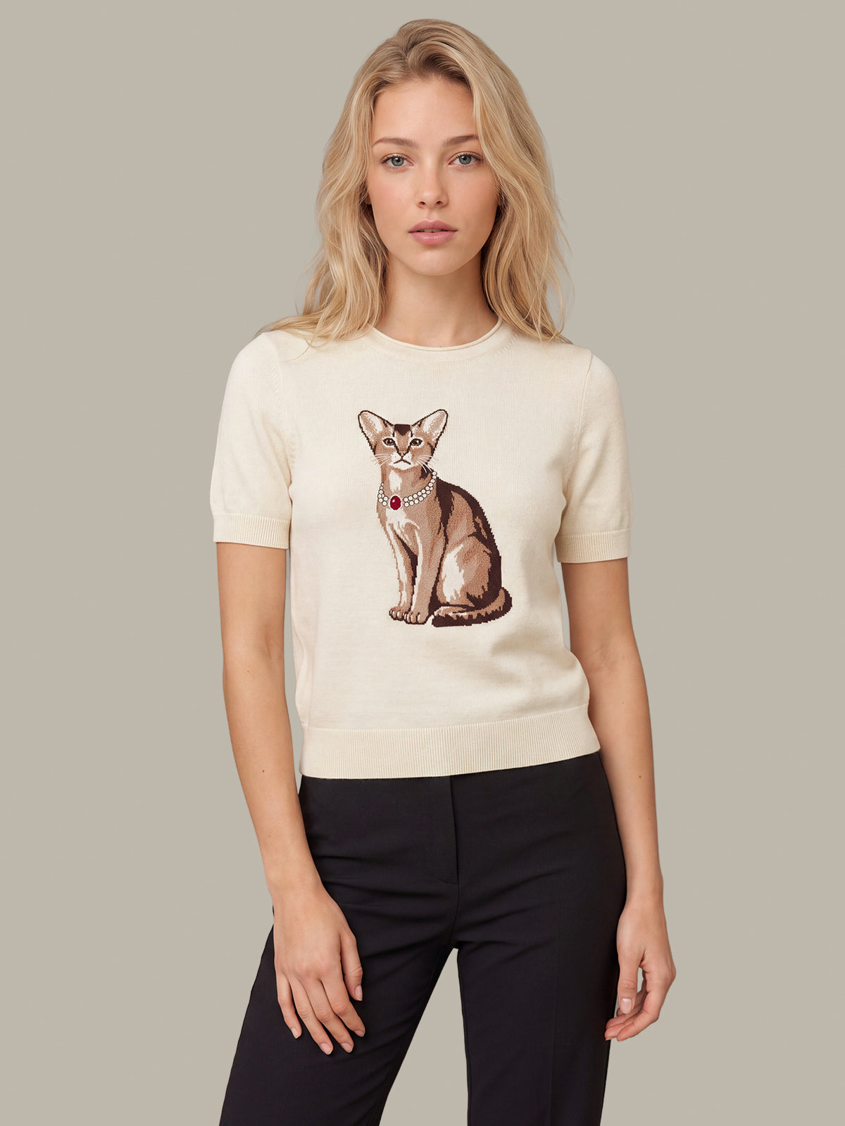 A woman modeling a cream-colored short-sleeve crewneck sweater featuring a detailed graphic of a sitting cat on the front, paired with black pants, set against a solid neutral gray background.