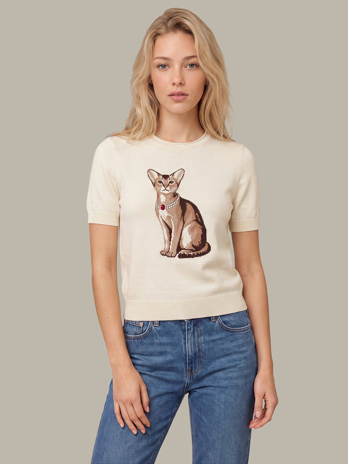 A woman wearing a cream-colored short-sleeve knit top with a detailed graphic of a sitting cat on the front, paired with blue jeans, set against a solid neutral gray background.