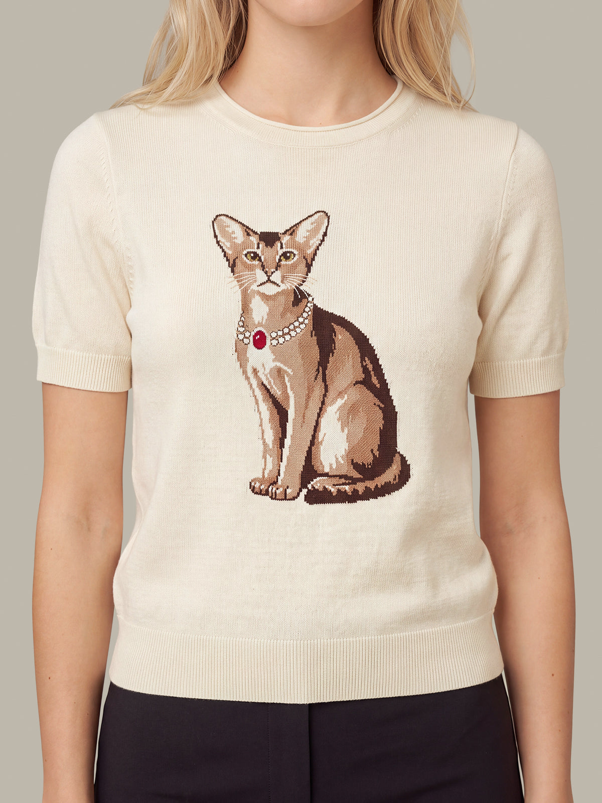 A woman wearing a cream-colored short-sleeve knit sweater with a detailed graphic of a brown cat wearing a pearl collar with a red jewel, paired with black pants.