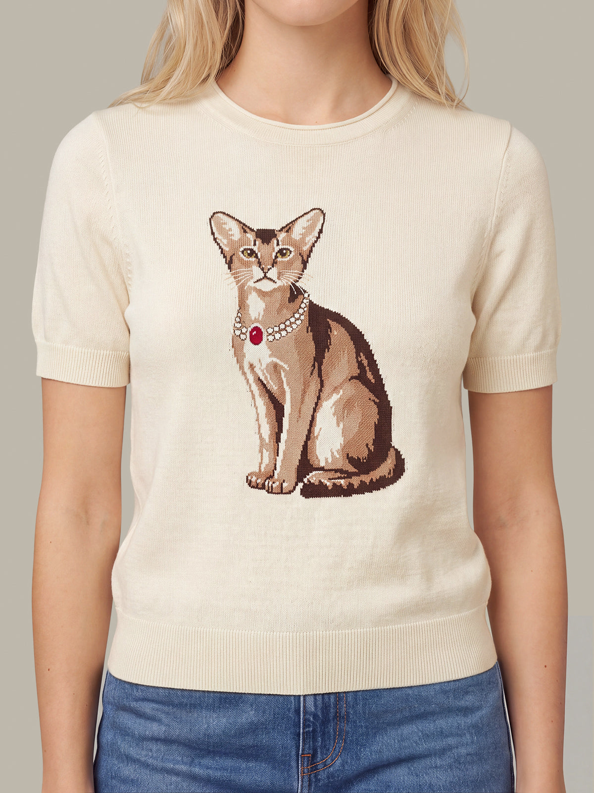 A woman wearing a cream-colored short-sleeve knit sweater with a detailed graphic of a brown cat wearing a pearl collar with a red jewel, paired with blue jeans.