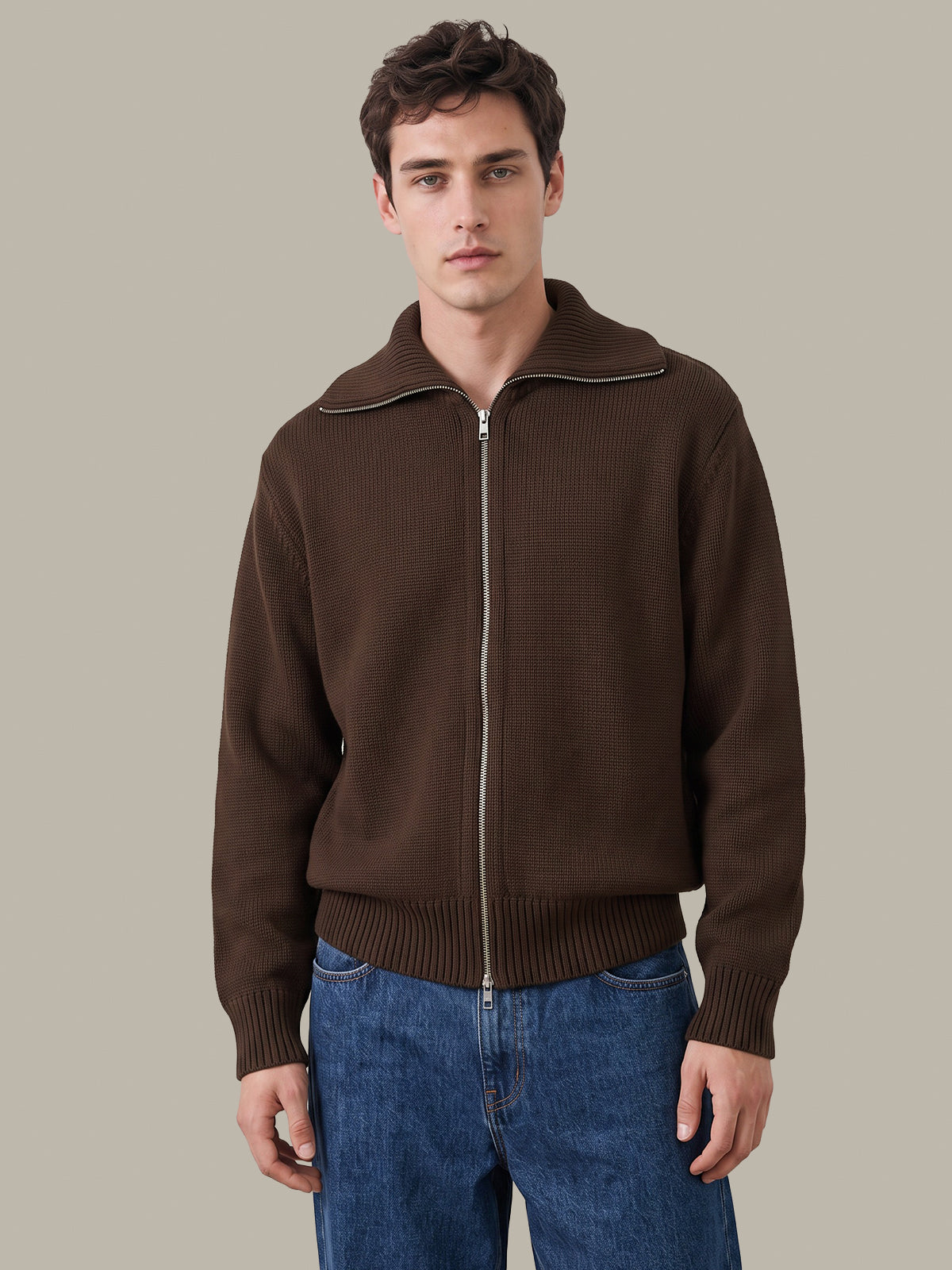 A man wearing a brown ribbed knit zip-up jacket with a stand collar and blue jeans, standing against a solid light gray background.