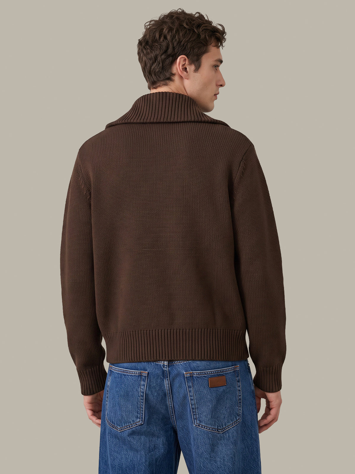 A man shown from the back wearing a dark brown ribbed knit sweater with a mock neck collar and blue jeans, set against a solid light gray background.
