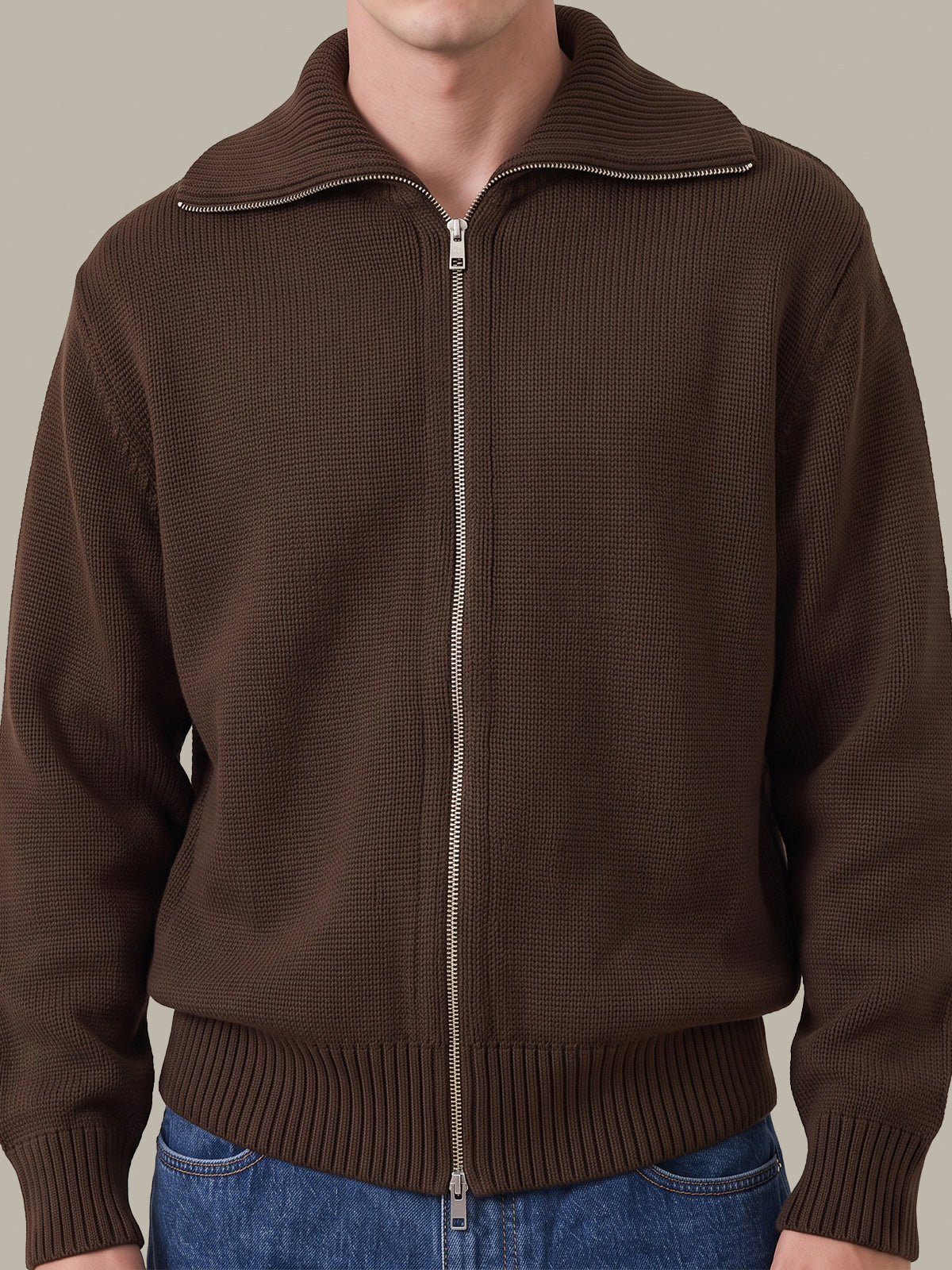 A man wearing a brown ribbed knit zip-up jacket with a stand collar and silver zipper, paired with blue jeans, against a solid light gray background.