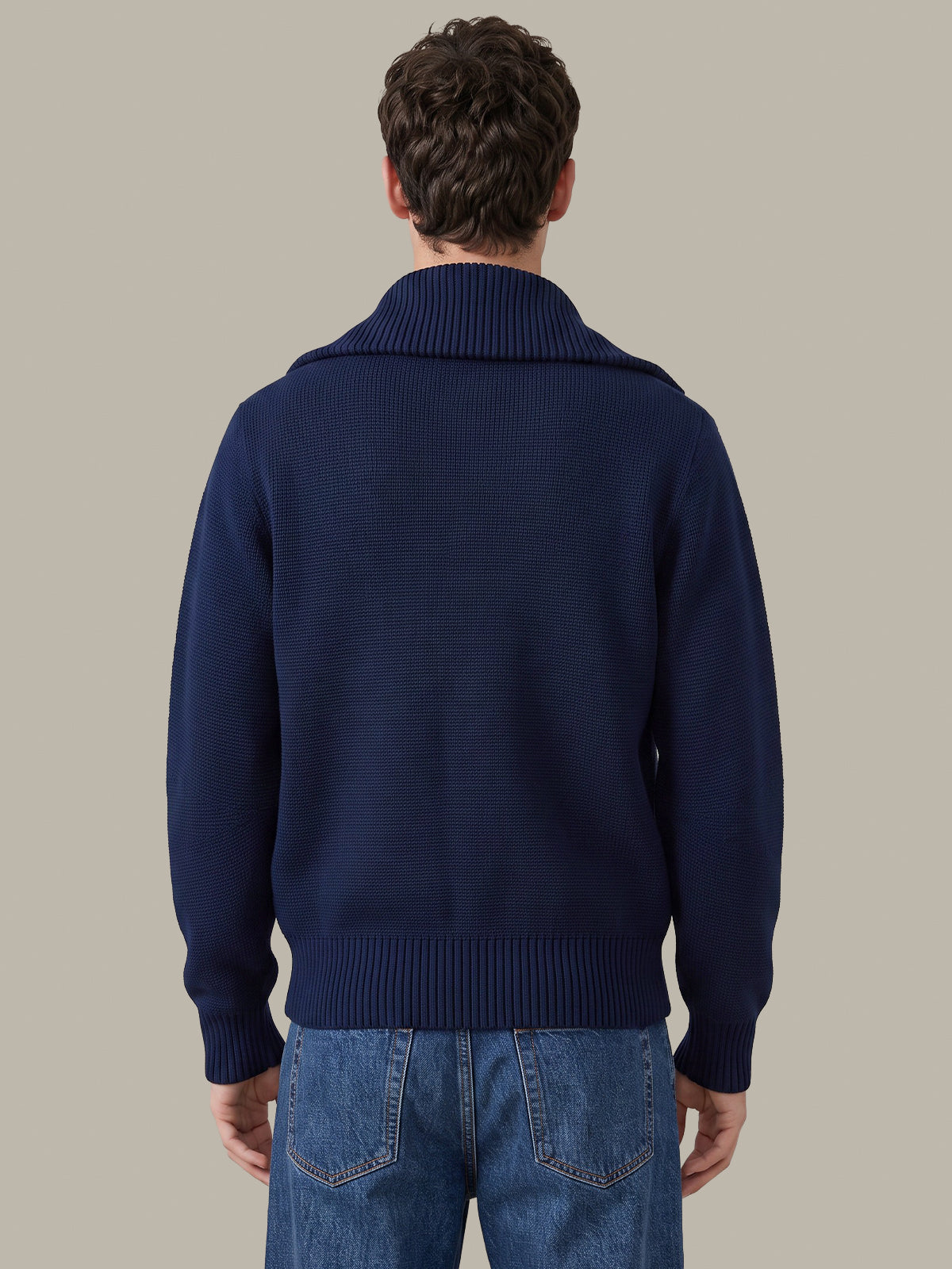 A man viewed from behind wearing a navy blue ribbed knit sweater and blue jeans, set against a solid light gray background.
