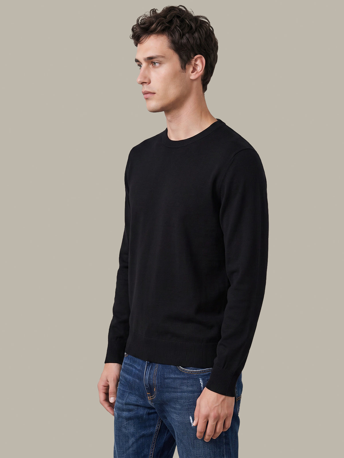 A man wearing a black long-sleeve crewneck sweater and blue jeans, standing in profile against a solid light gray background.