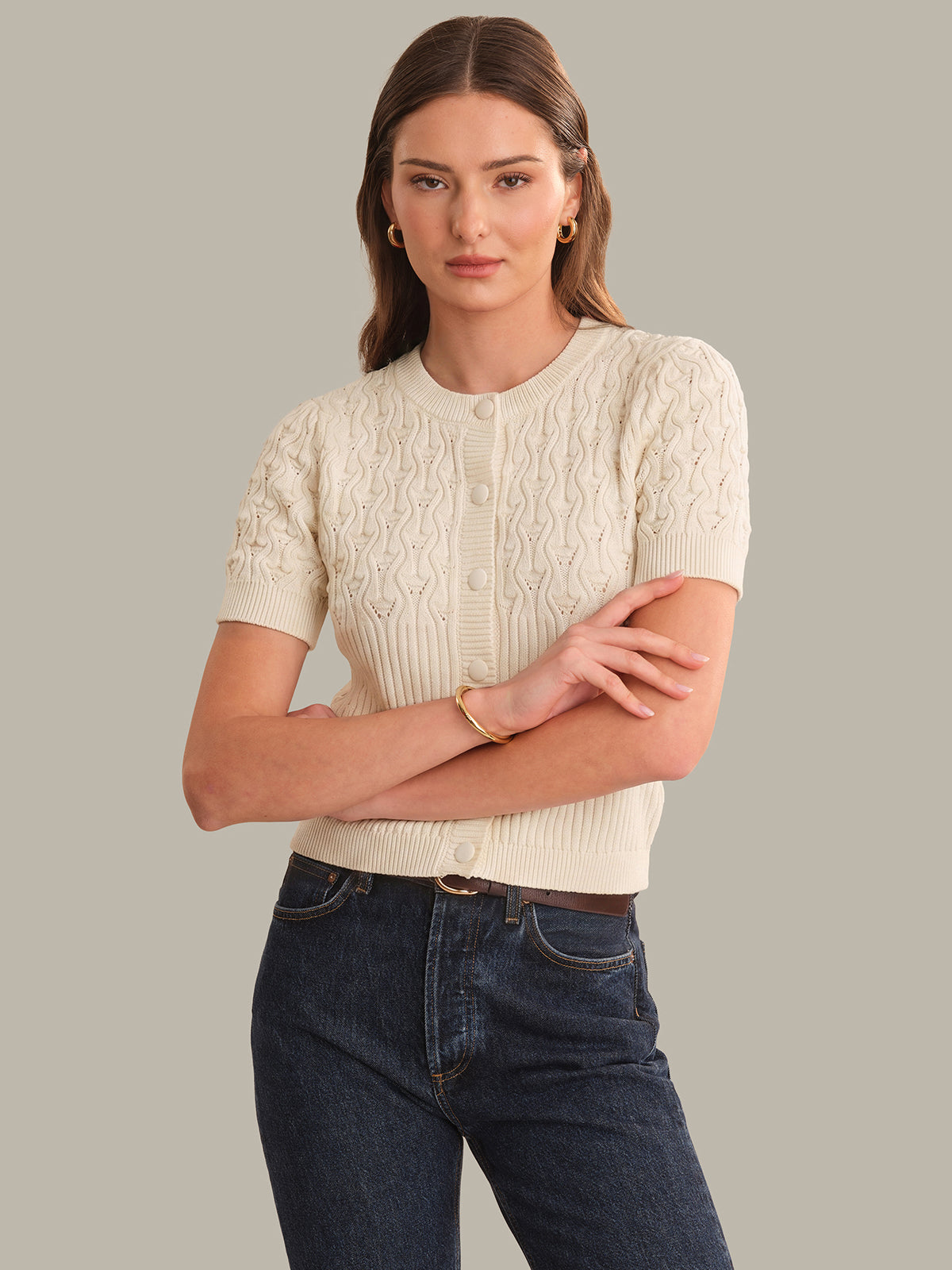 Woman wearing a cream knitted cardigan and dark jeans against a gray background